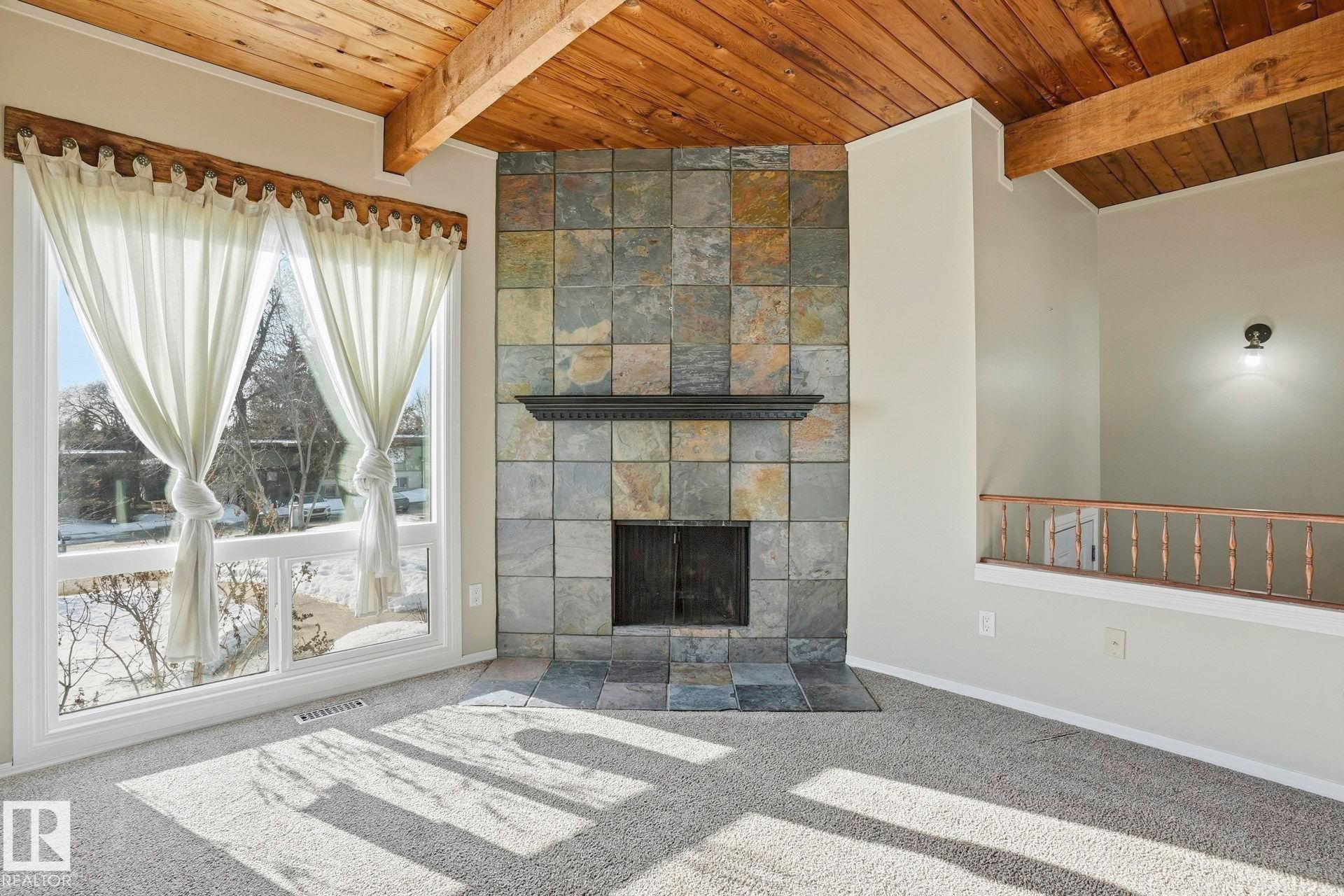 Unfurnished living room featuring a wooden ceiling with exposed beams, a tile fireplace, and carpet floors - Edmonton, AB - Indoor Photo Showing Other Room With Fireplace