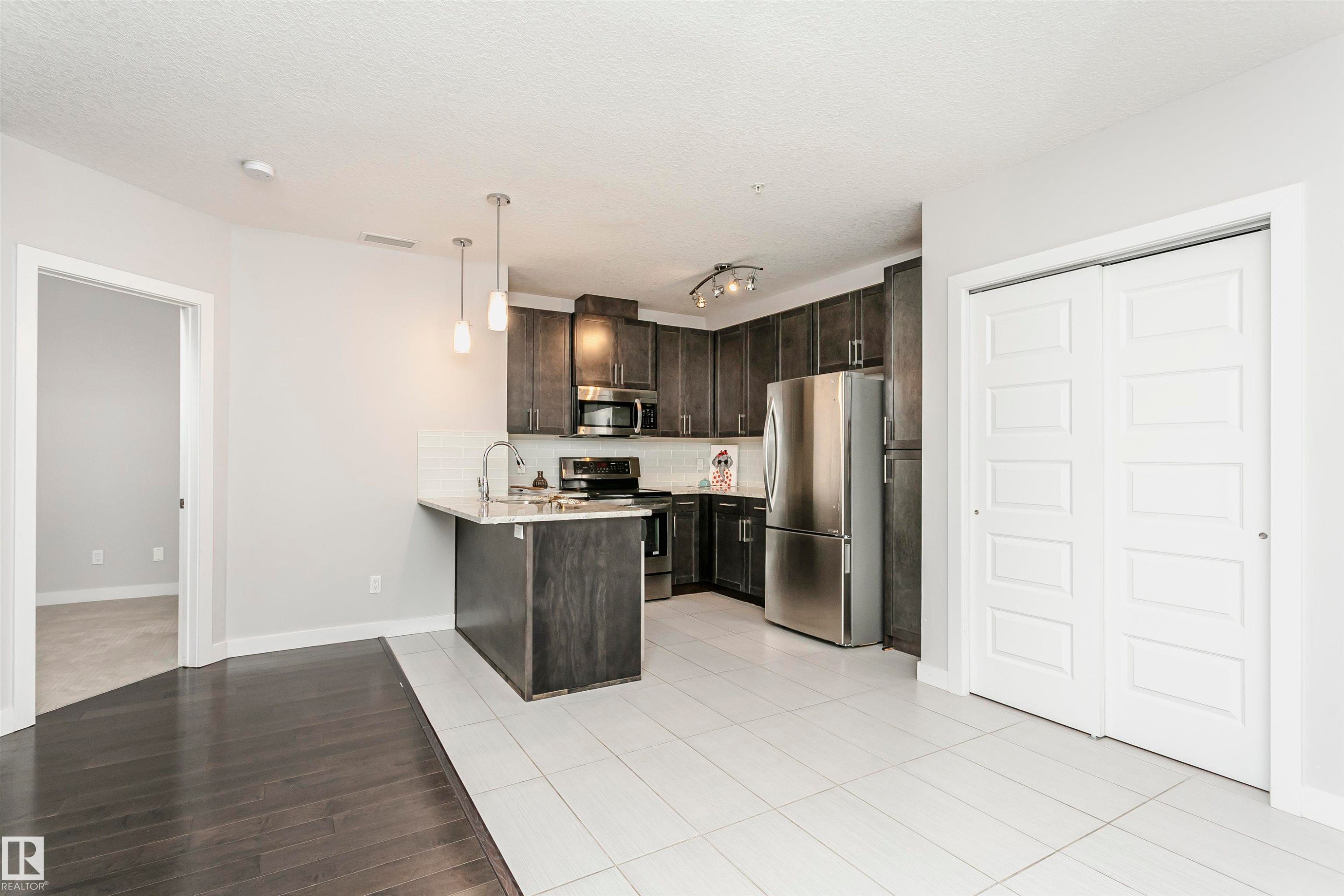 Kitchen featuring stainless steel appliances, dark wood finish cabinets, a peninsula, tasteful backsplash, and pendant lighting - 332 5151 Windermere Boulevard, Edmonton, AB - Indoor Photo Showing Kitchen