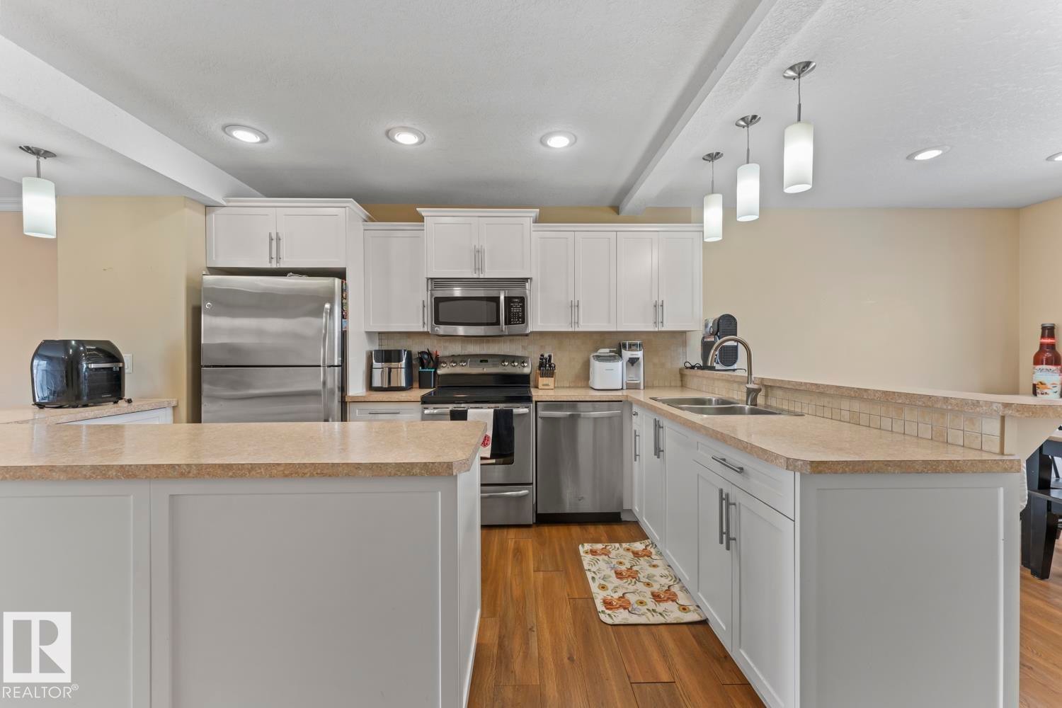 Kitchen featuring pendant lighting, a peninsula, white cabinetry, stainless steel appliances, and beamed ceiling - 7 1001 7 Avenue, Cold Lake, AB - Indoor Photo Showing Kitchen With Double Sink