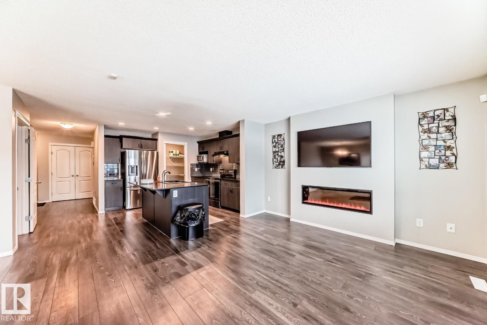 Kitchen with a kitchen bar, stainless steel appliances, an island with sink, a glass covered fireplace, and dark wood finished floors - 2823 Duke Crescent, Edmonton, AB - Indoor Photo Showing Living Room With Fireplace