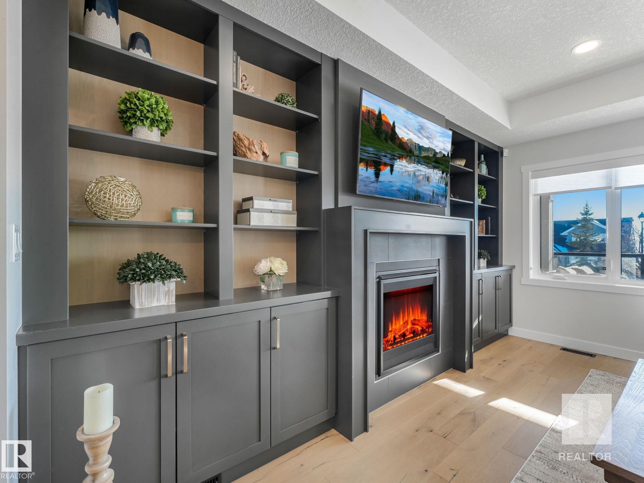 Living room featuring built in shelves, light wood-style floors, a textured ceiling, and a warm lit fireplace - 209 55101 Ste. Anne Trail, Rural Lac Ste. Anne County, AB - Indoor With Fireplace