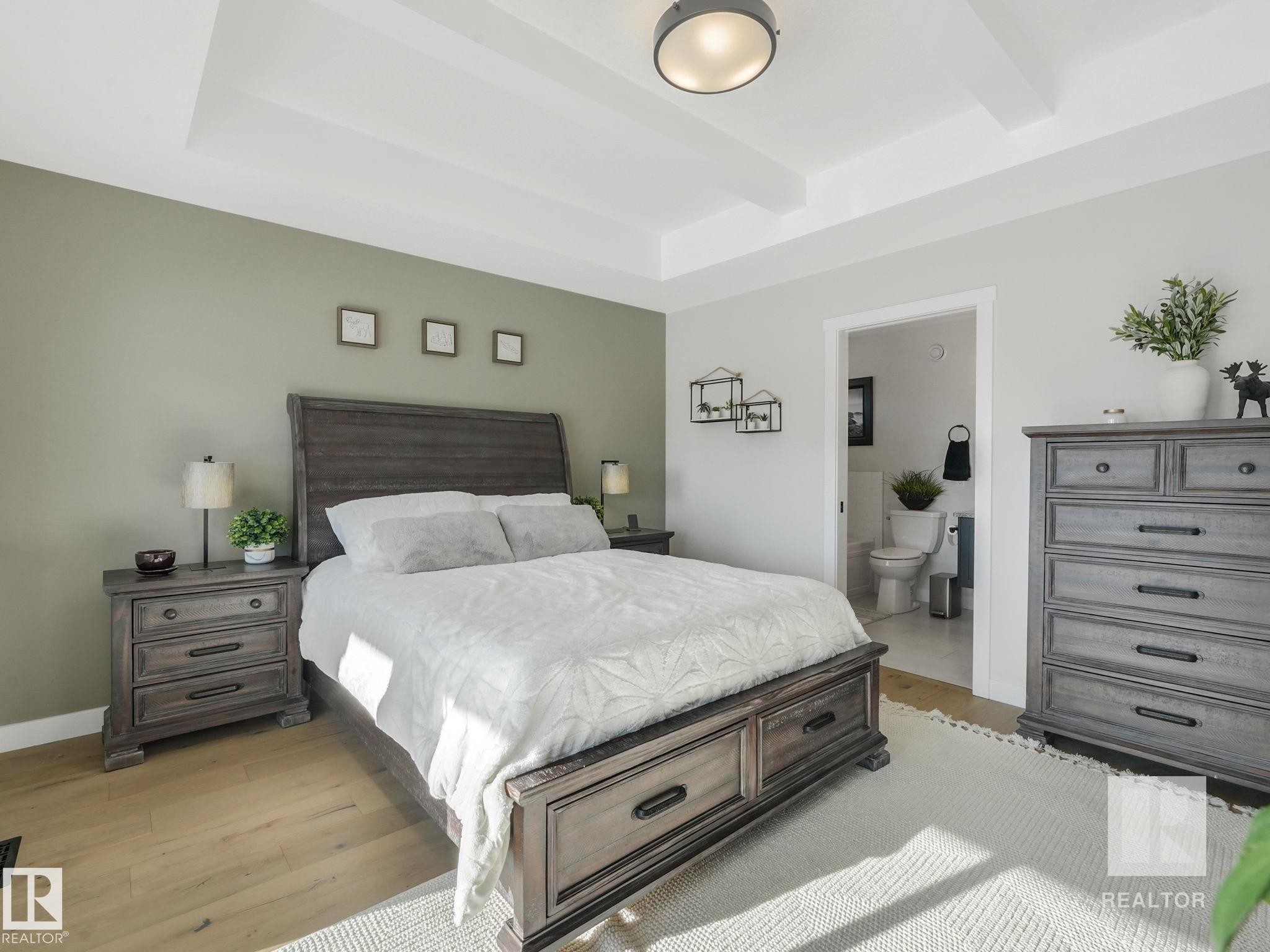 Bedroom with light wood-style floors, beamed ceiling, a raised ceiling, and ensuite bath - 209 55101 Ste. Anne Trail, Rural Lac Ste. Anne County, AB - Indoor Photo Showing Bedroom