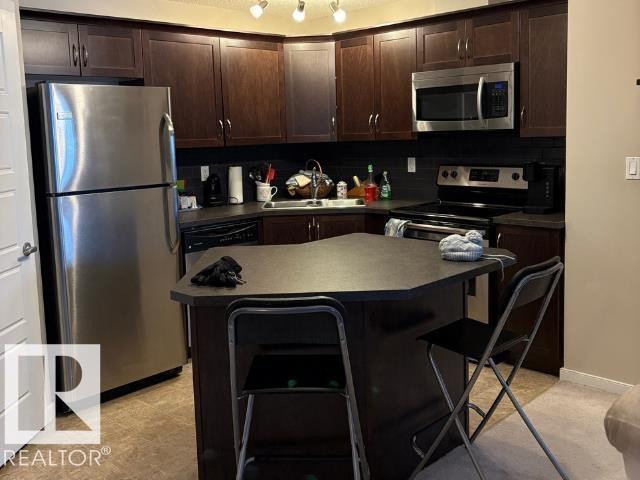 Kitchen with dark wood finish cabinets, a kitchen bar, stainless steel appliances, dark countertops, and a center island - 118 396 Silverberry Road Nw, Edmonton, AB - Indoor Photo Showing Kitchen