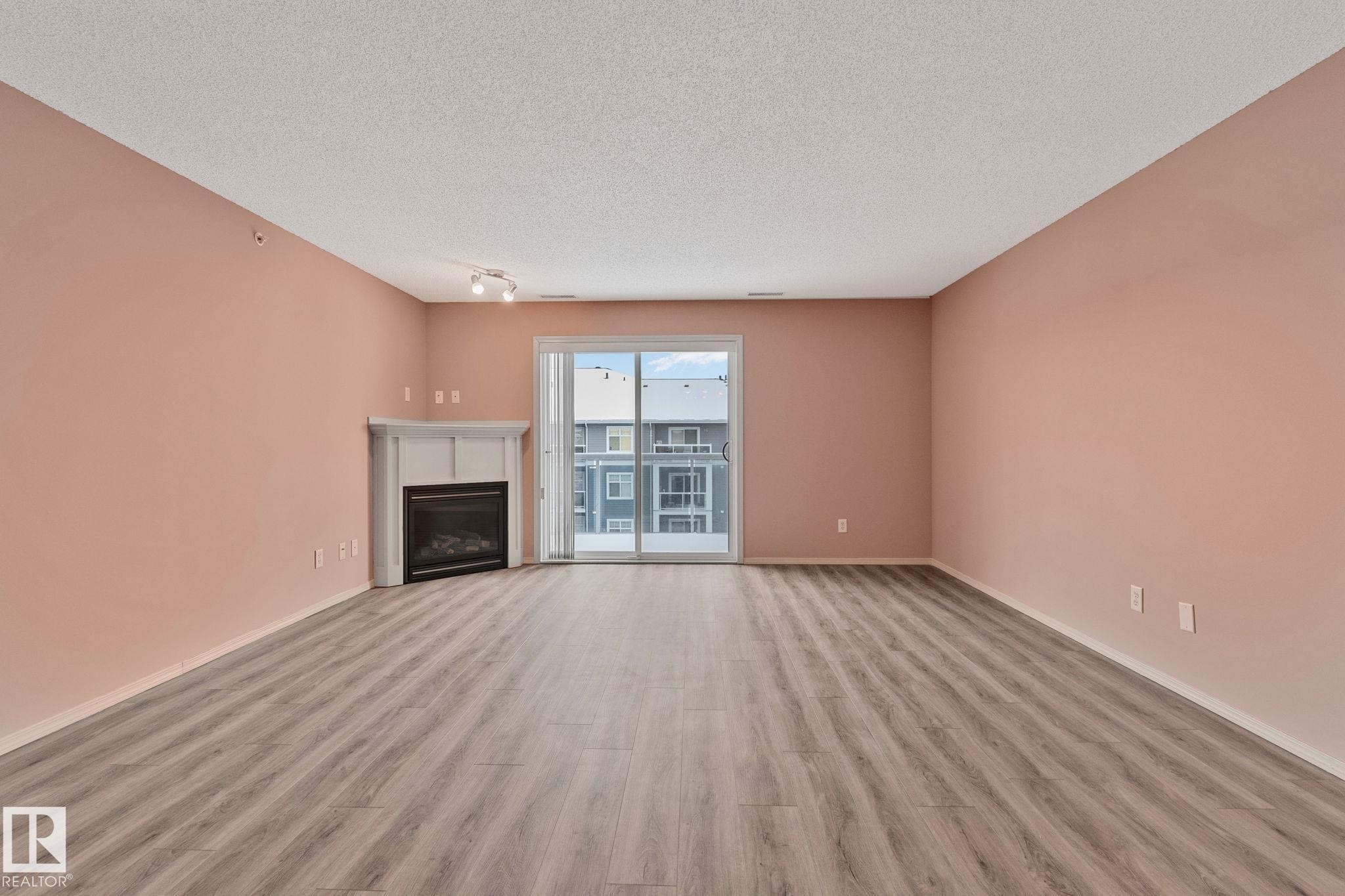Unfurnished living room with light wood-type flooring, a fireplace, and a textured ceiling - 406 16035 132 Street Nw, Edmonton, AB - Indoor Photo Showing Other Room With Fireplace