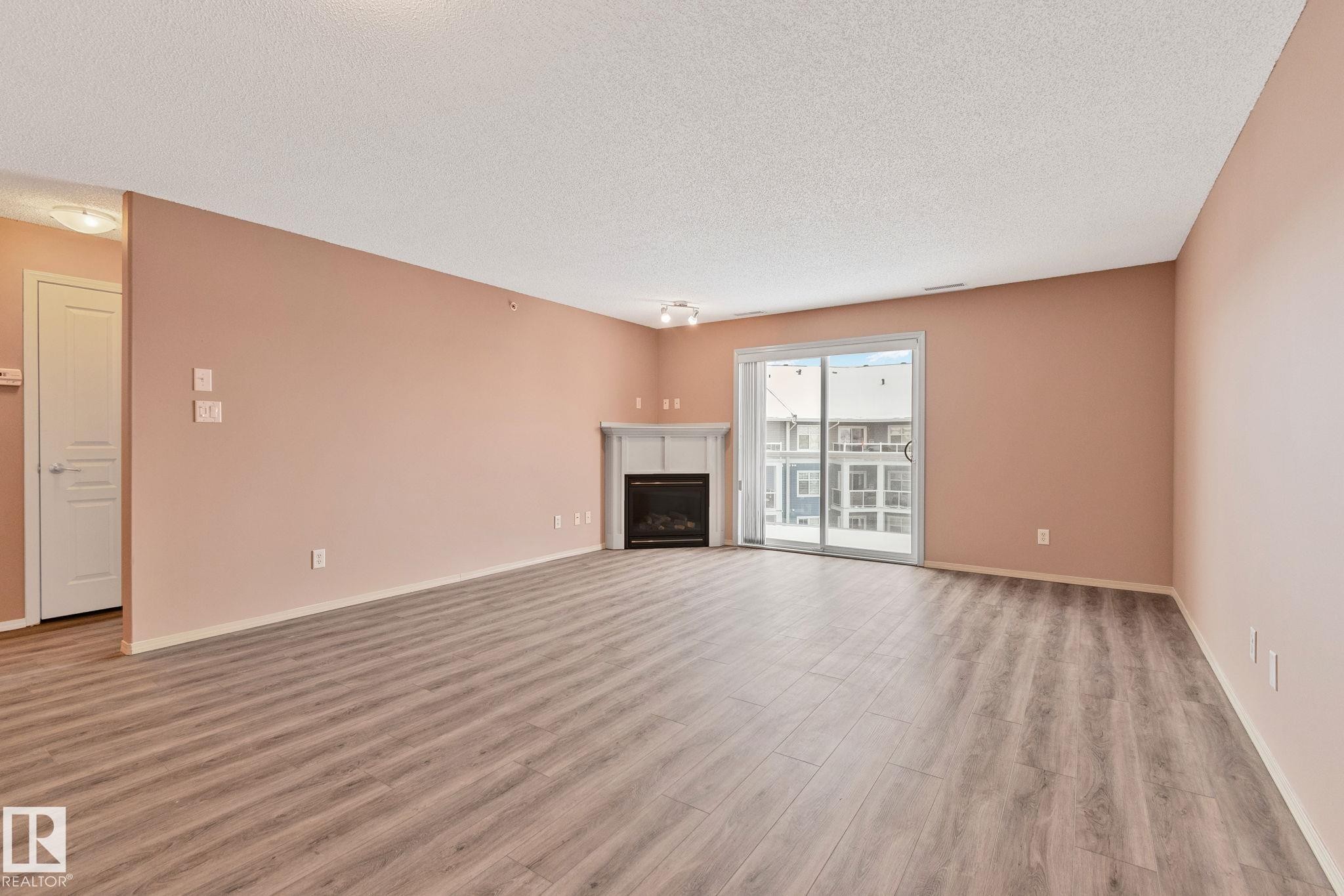 Unfurnished living room with light wood-type flooring, a fireplace, and a textured ceiling - 406 16035 132 Street Nw, Edmonton, AB - Indoor Photo Showing Living Room With Fireplace