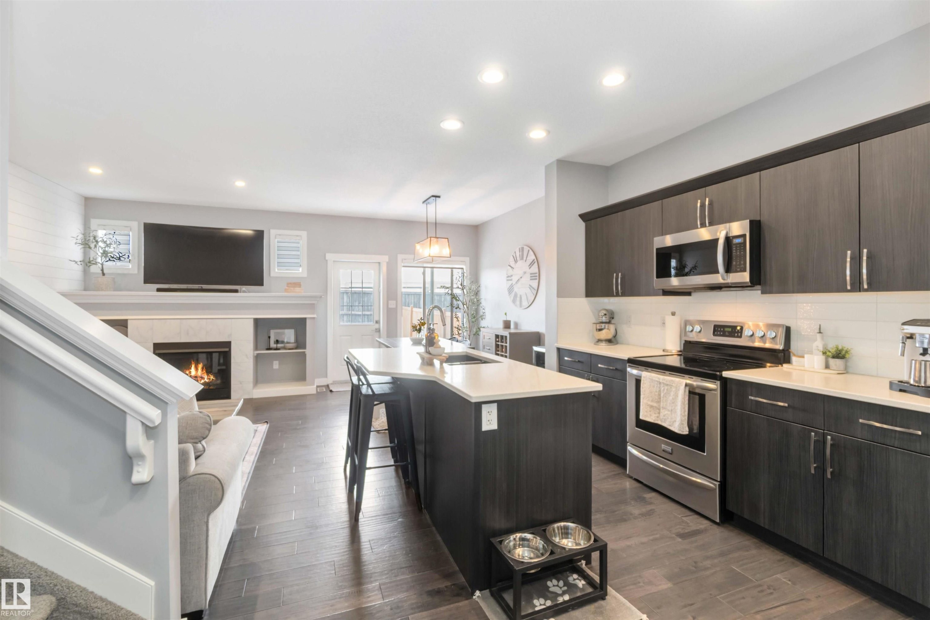Kitchen featuring stainless steel appliances, a tiled fireplace, dark wood finish cabinetry, a breakfast bar, and a kitchen island with sink - 17143 38 Street, Edmonton, AB - Indoor Photo Showing Kitchen With Fireplace With Double Sink With Upgraded Kitchen