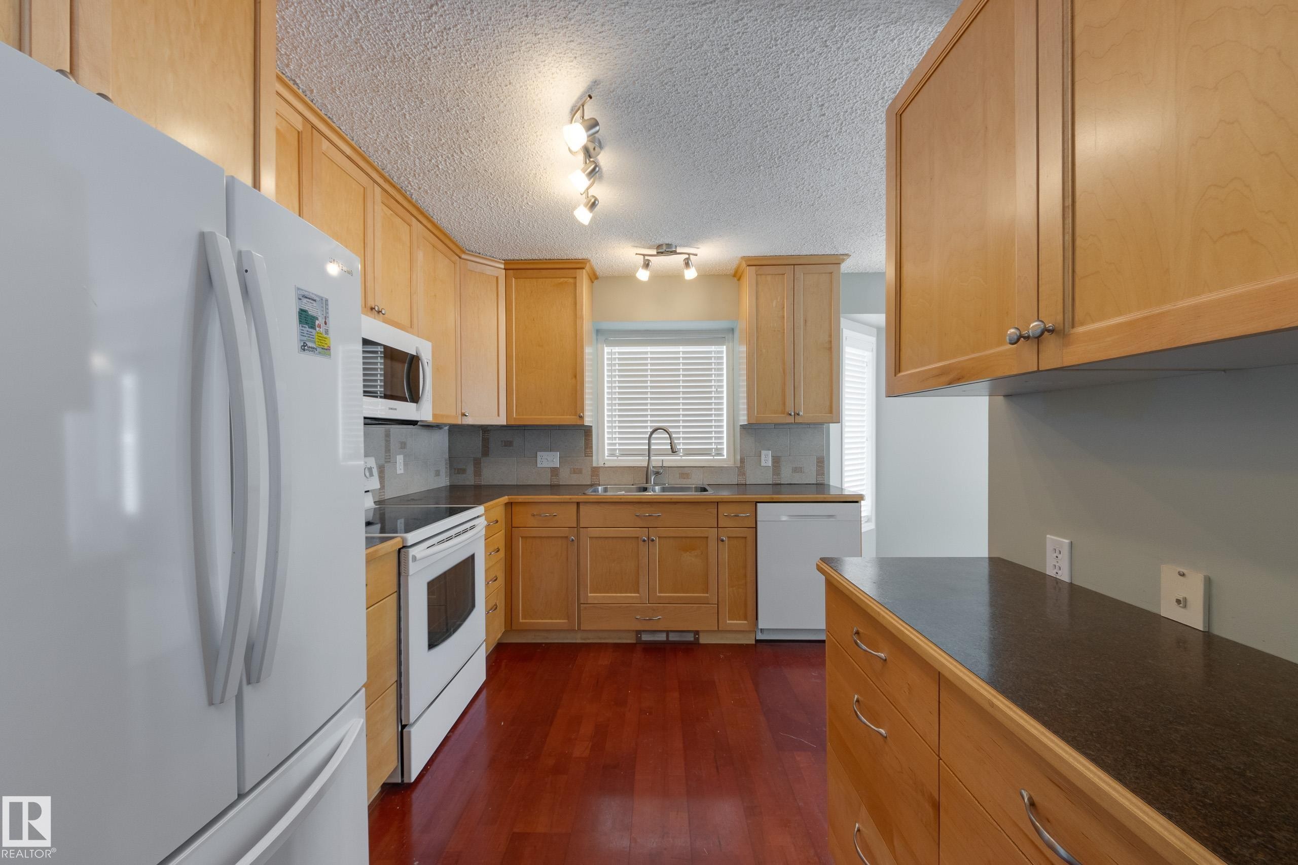 Kitchen with white appliances, dark countertops, decorative backsplash, dark wood-type flooring, and a textured ceiling - 9916 178 Avenue, Edmonton, AB - Indoor Photo Showing Kitchen With Double Sink