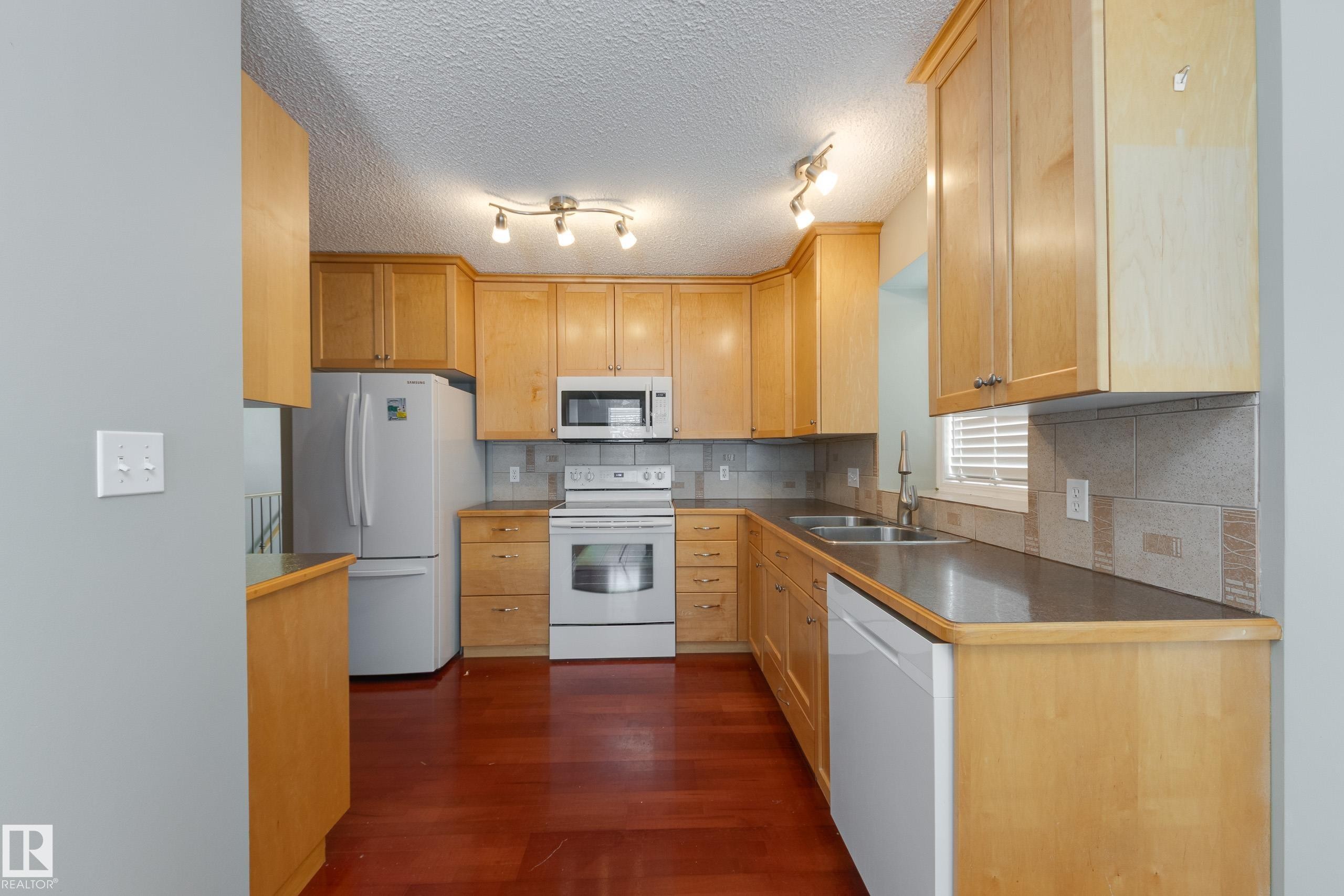 Kitchen with white appliances, a textured ceiling, dark wood-type flooring, backsplash, and light wood finish cabinets - 9916 178 Avenue, Edmonton, AB - Indoor Photo Showing Kitchen With Double Sink