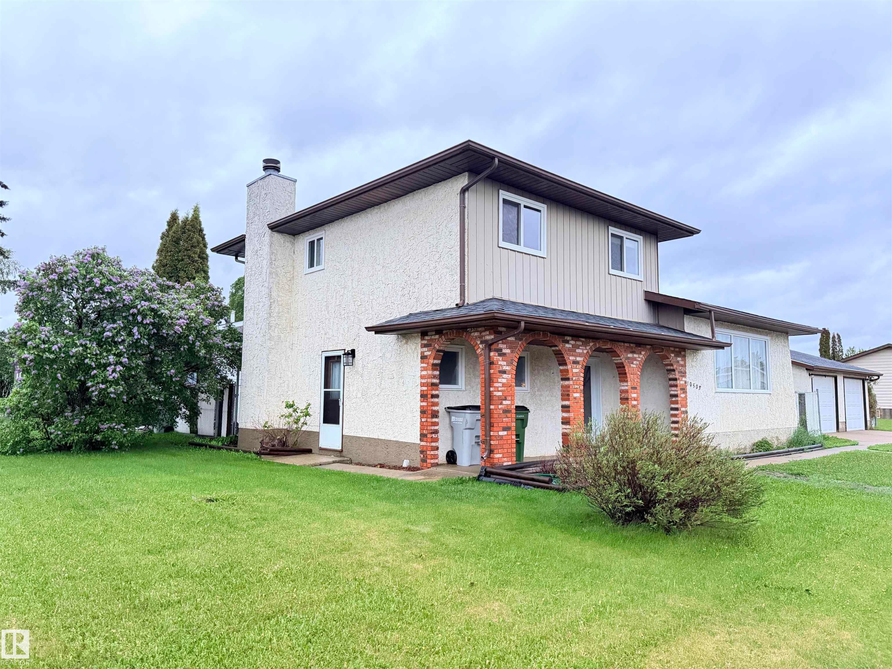 Exterior showcasing a combination of stucco and vertical siding, a covered arched brick entryway, multiple windows, an attached garage, and a lush green lawn - 10503 110 Street, Westlock, AB