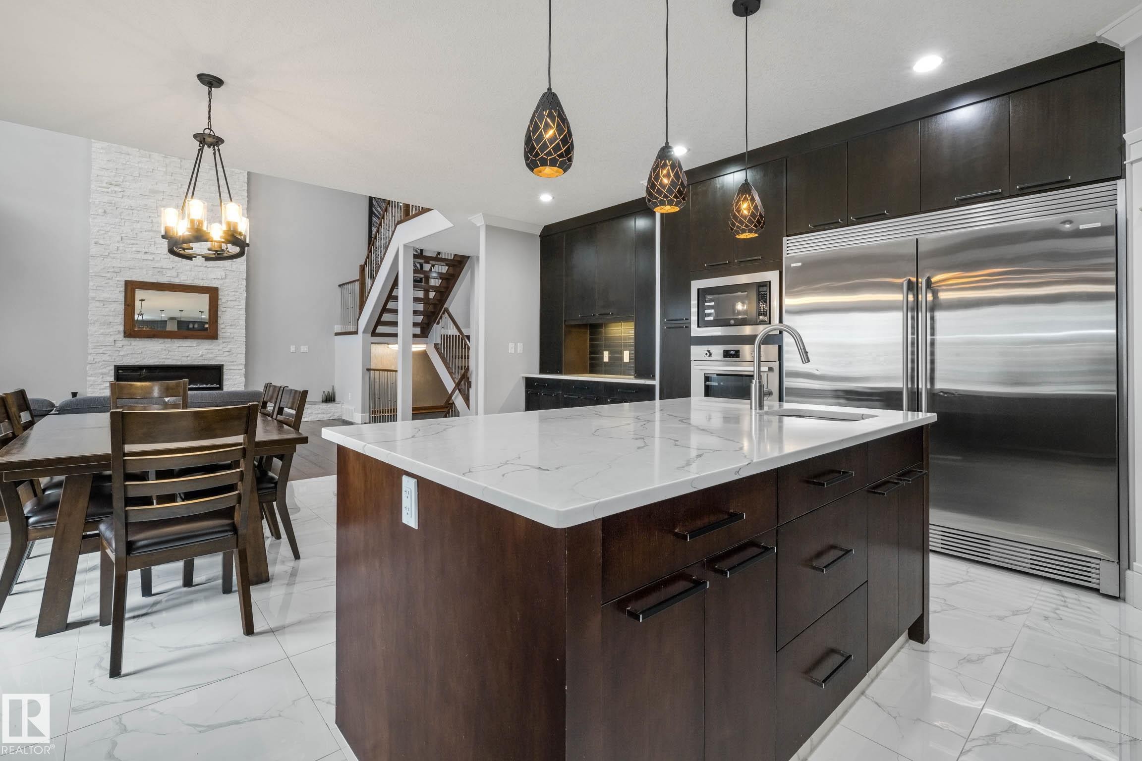 Kitchen with built in appliances, a kitchen island with sink, dark wood finish cabinetry, a stone fireplace, and light stone countertops - 4086 Whispering River Drive, Edmonton, AB - Indoor Photo Showing Kitchen With Stainless Steel Kitchen With Upgraded Kitchen