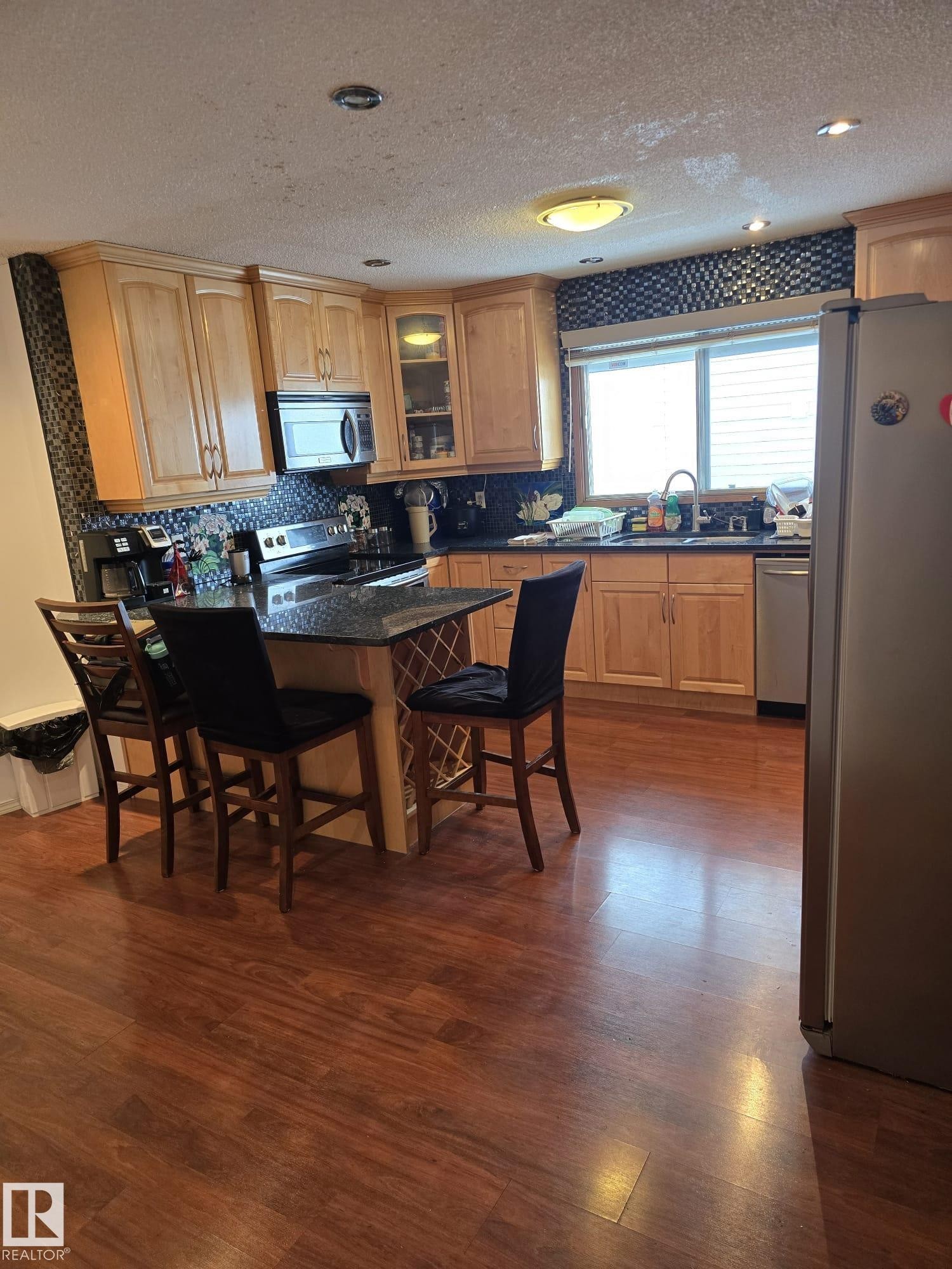 Kitchen with a kitchen breakfast bar, stainless steel appliances, a textured ceiling, glass insert cabinets, and dark wood finished floors - 11927 85 Street Nw, Edmonton, AB - Indoor Photo Showing Kitchen With Double Sink