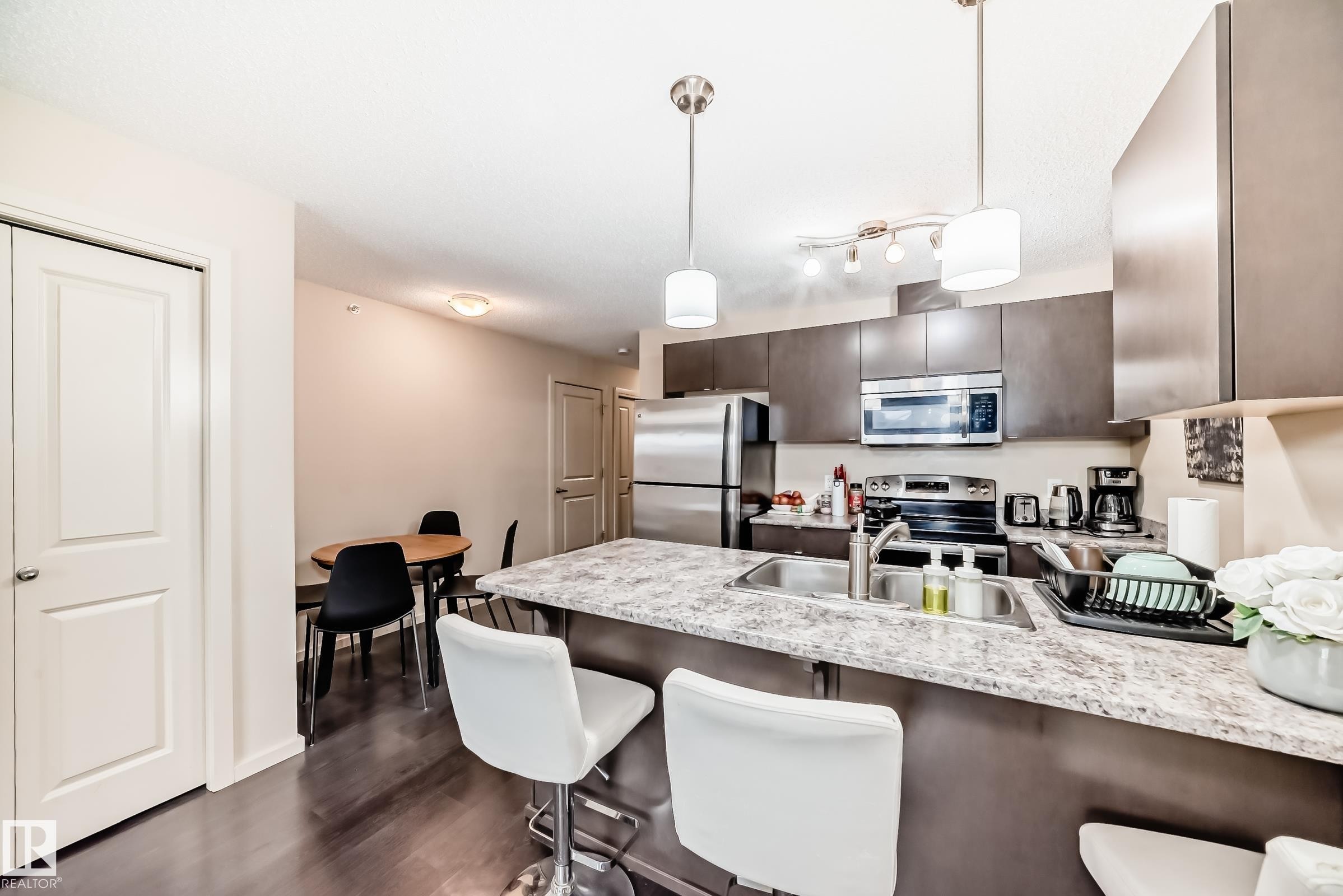 Kitchen with a kitchen bar, a peninsula, dark wood finish cabinetry, stainless steel appliances, and hanging light fixtures - 403 340 Windermere Road, Edmonton, AB - Indoor Photo Showing Kitchen With Stainless Steel Kitchen With Upgraded Kitchen
