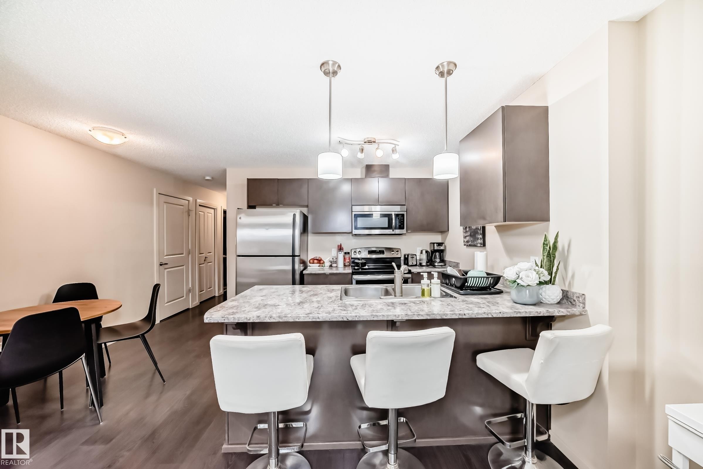 Kitchen featuring a peninsula, a kitchen breakfast bar, stainless steel appliances, pendant lighting, and dark wood finish cabinetry - 403 340 Windermere Road, Edmonton, AB - Indoor