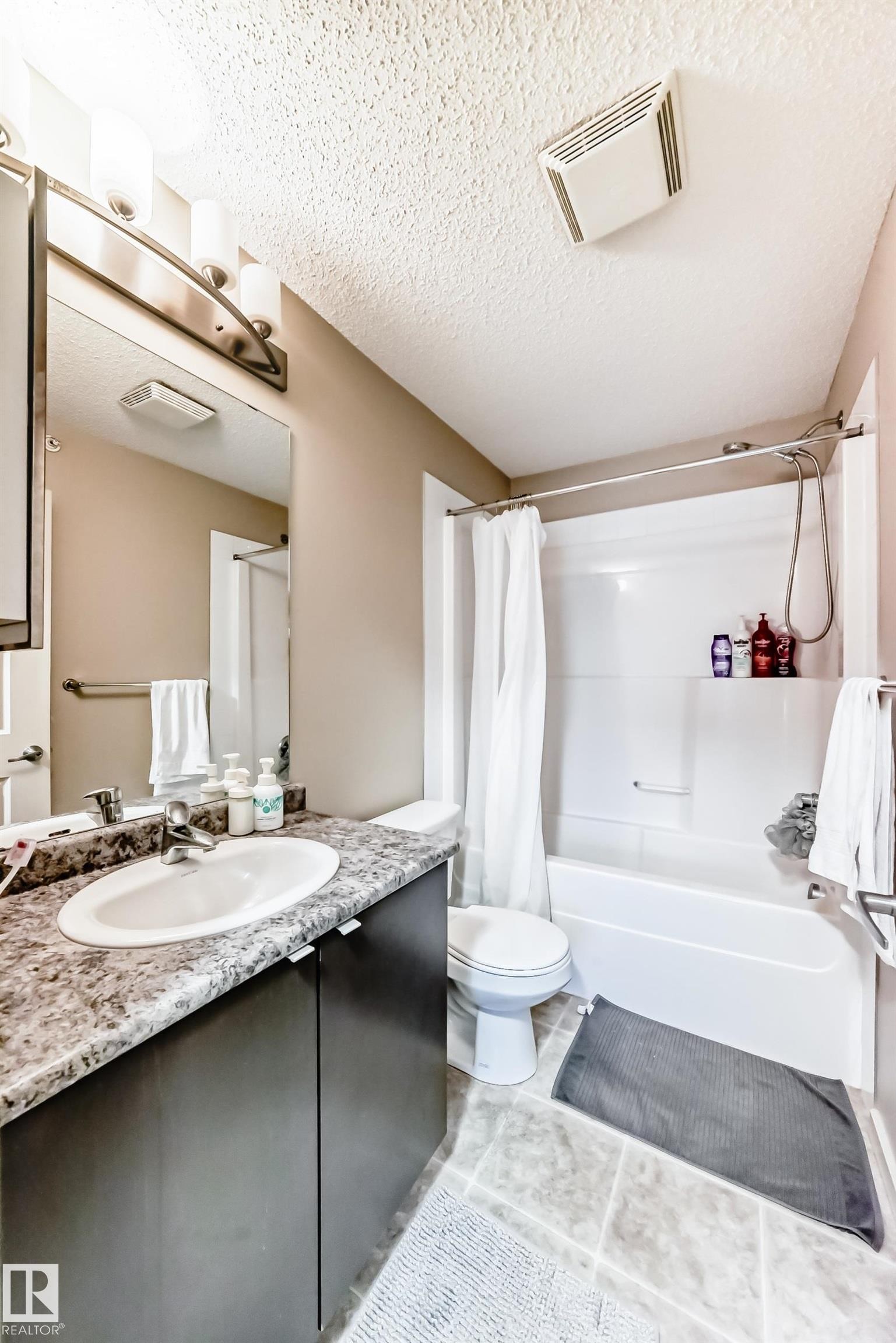 Full bath featuring vanity, shower / bath combination with curtain, a textured ceiling, and light tile patterned floors - 403 340 Windermere Road, Edmonton, AB - Indoor Photo Showing Bathroom