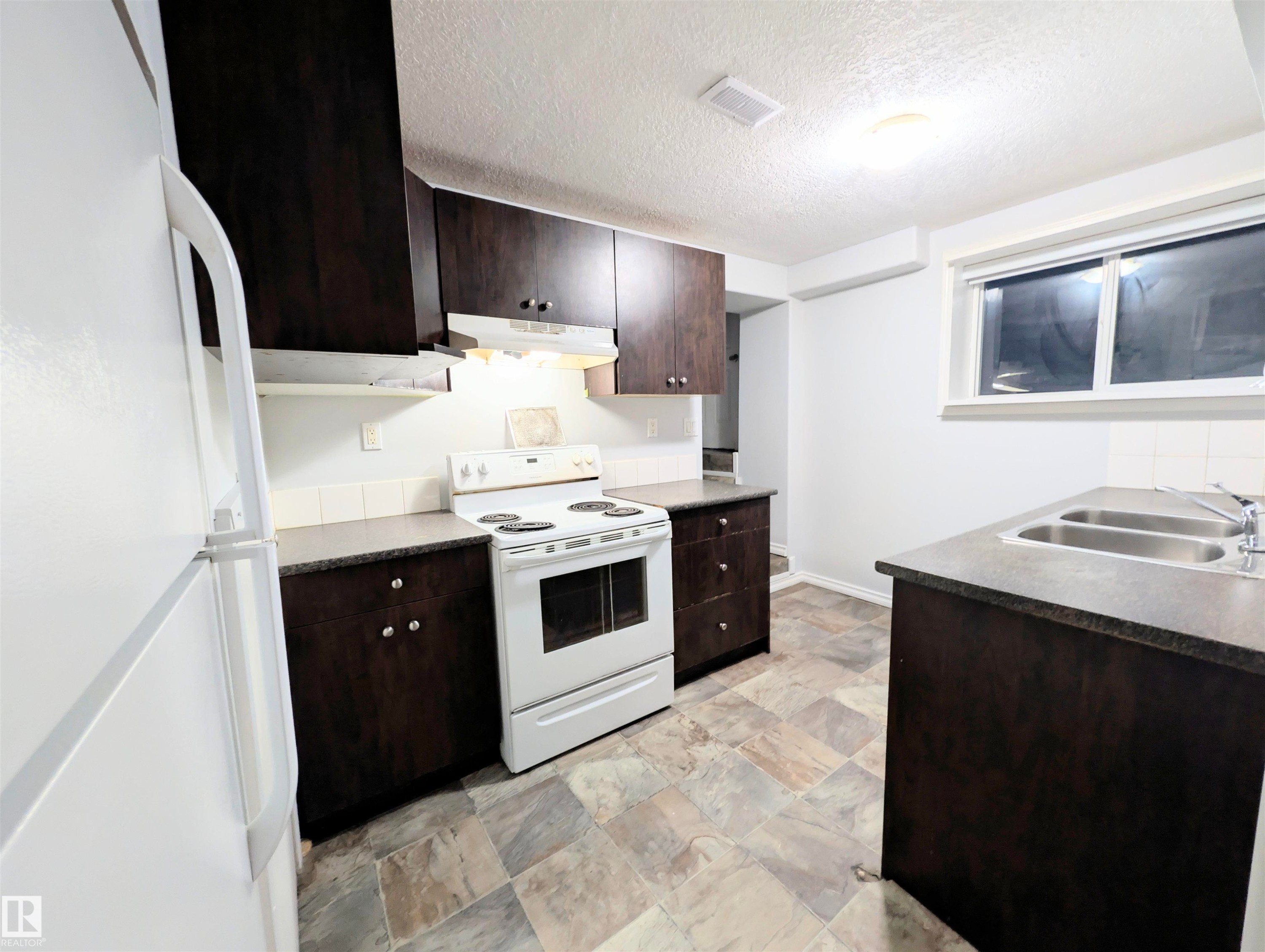 Kitchen featuring white appliances, dark wood finish cabinets, dark countertops, a textured ceiling, and stone finish floors - 3307 24 Avenue, Edmonton, AB - Indoor Photo Showing Kitchen With Double Sink