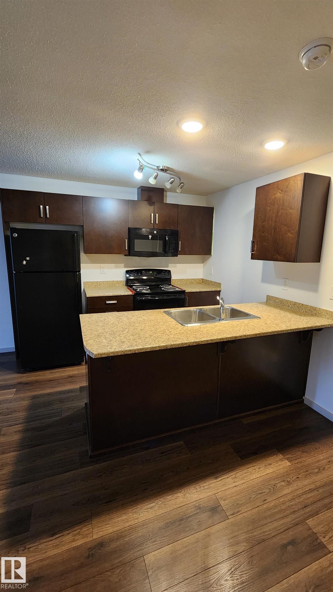 Kitchen with black appliances, dark wood finish cabinetry, a textured ceiling, a peninsula, and dark wood finished floors - 413 5521 7 Avenue, Edmonton, AB - Indoor Photo Showing Kitchen