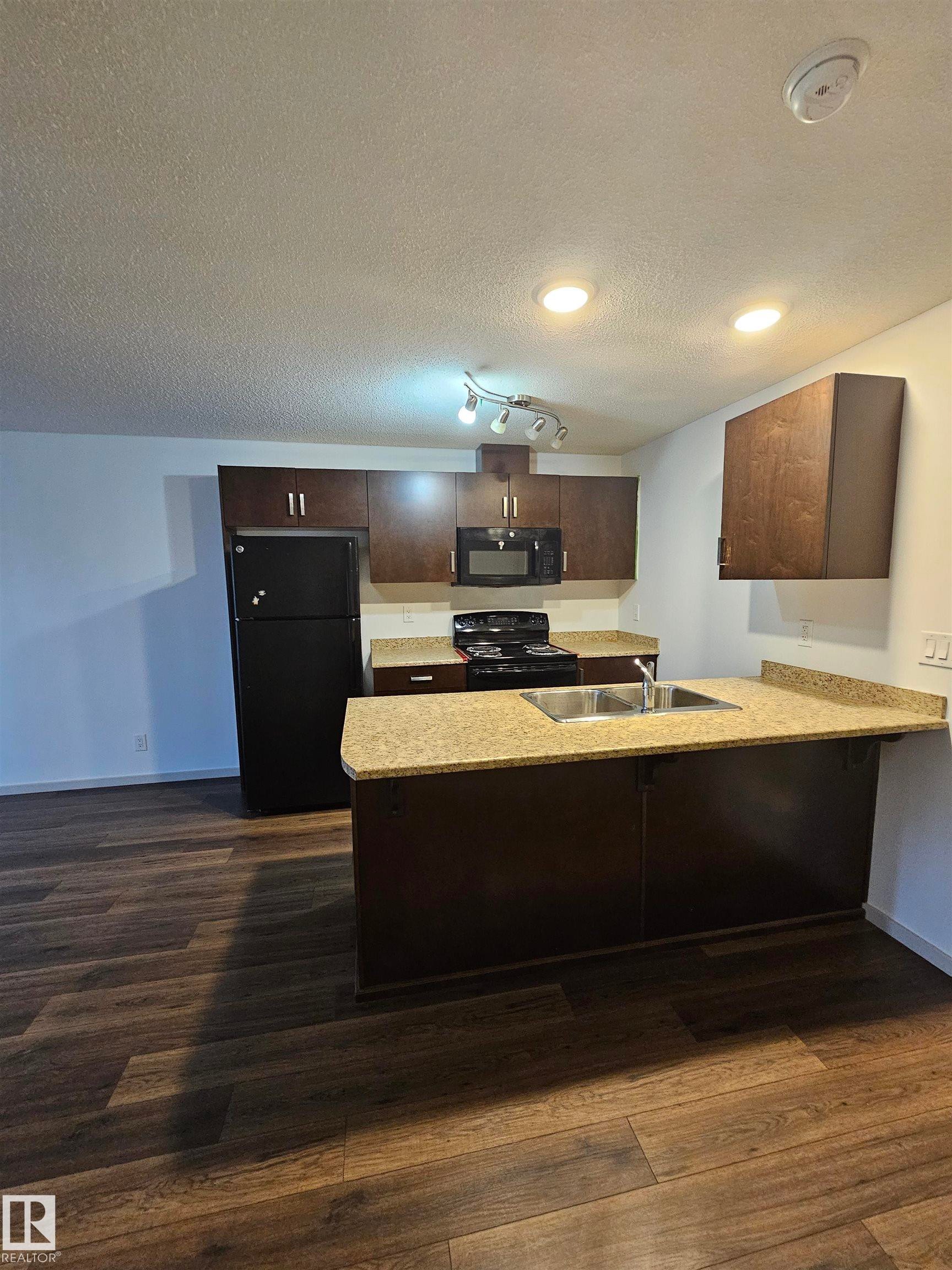 Kitchen featuring a peninsula, dark wood finish cabinets, black appliances, light countertops, and a textured ceiling - 413 5521 7 Avenue, Edmonton, AB - Indoor Photo Showing Kitchen With Double Sink