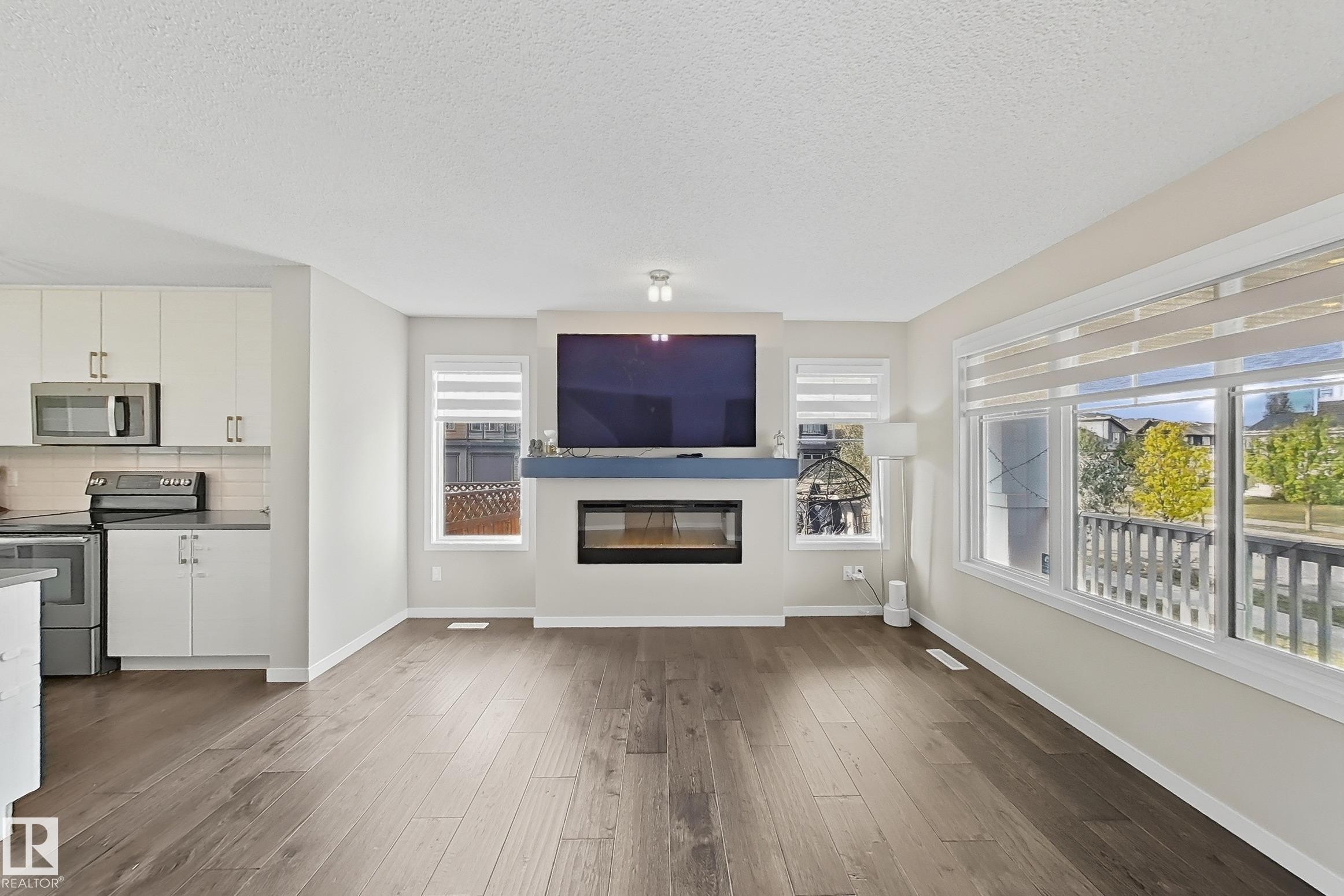 Unfurnished living room featuring wood-type flooring, a glass covered fireplace, plenty of natural light, and a textured ceiling - 4303 Prowse Link, Edmonton, AB - Indoor With Fireplace