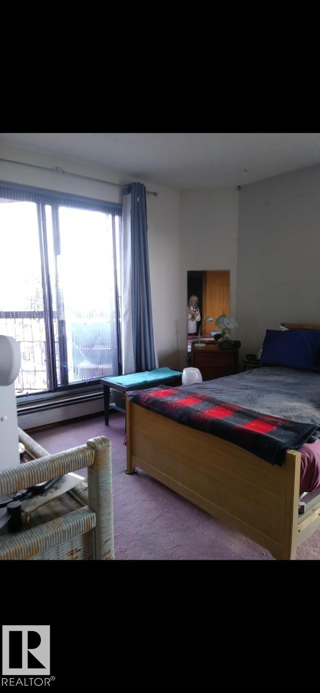 This room features a large window with vertical blinds and curtains, a baseboard heater, and carpeting - 401 10743 107 Street Nw, Edmonton, AB - Indoor Photo Showing Bedroom