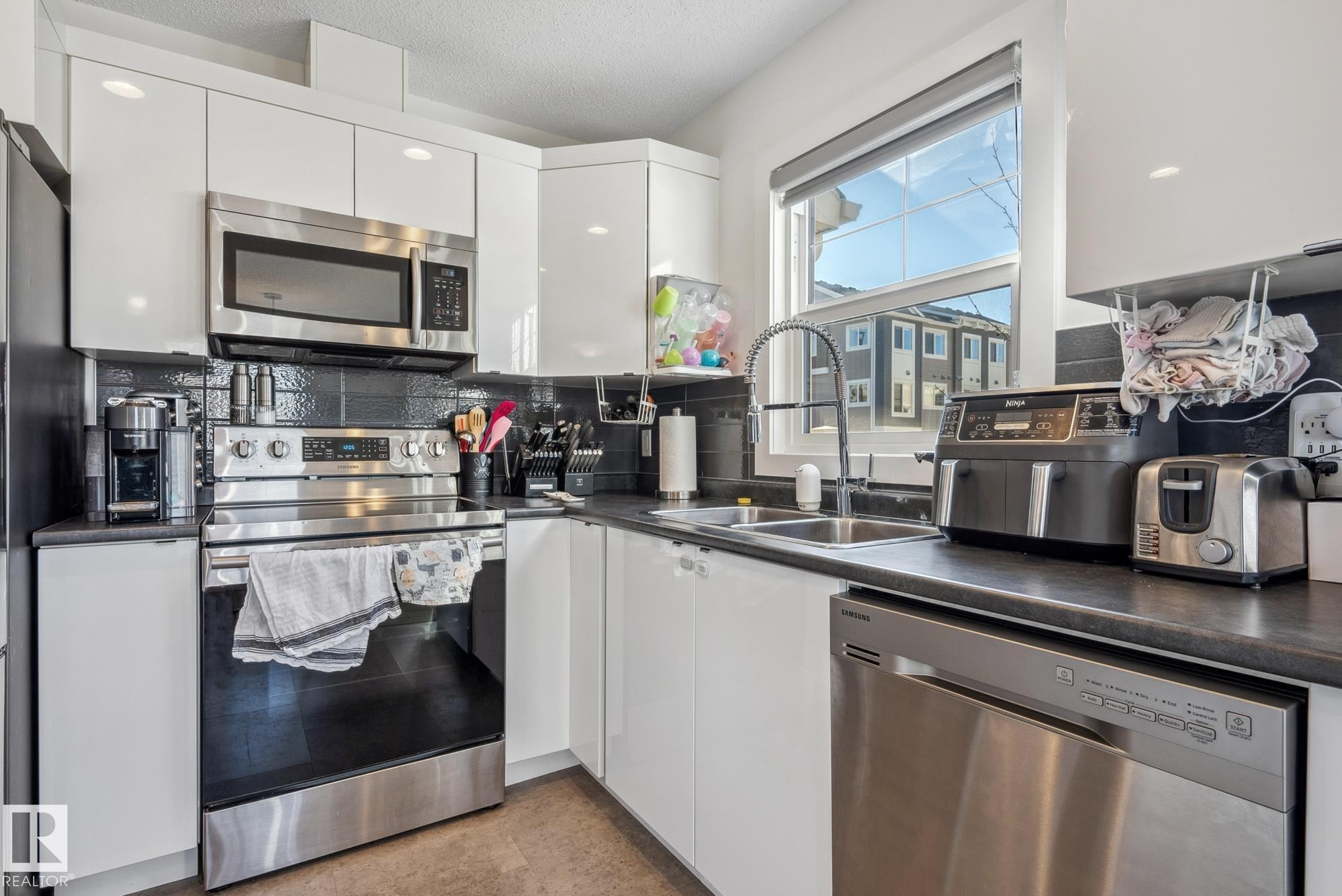 Kitchen featuring stainless steel appliances, dark countertops, white cabinetry, and a textured ceiling - 76 330 Bulyea Road, Edmonton, AB - Indoor Photo Showing Kitchen With Double Sink