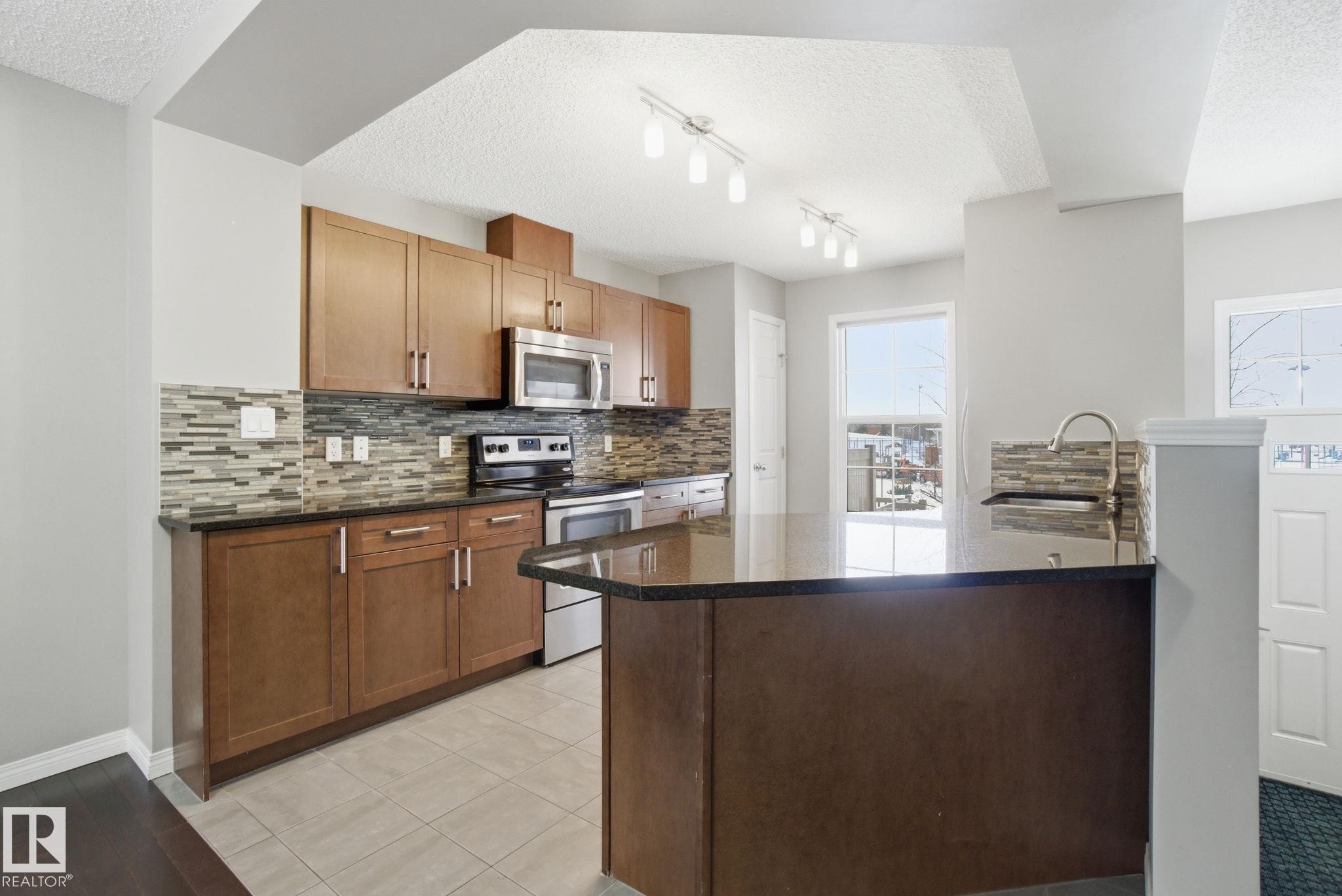 Kitchen with stainless steel appliances, a peninsula, dark stone counters, a textured ceiling, and decorative backsplash - 71 4029 Orchards Drive, Edmonton, AB - Indoor Photo Showing Kitchen
