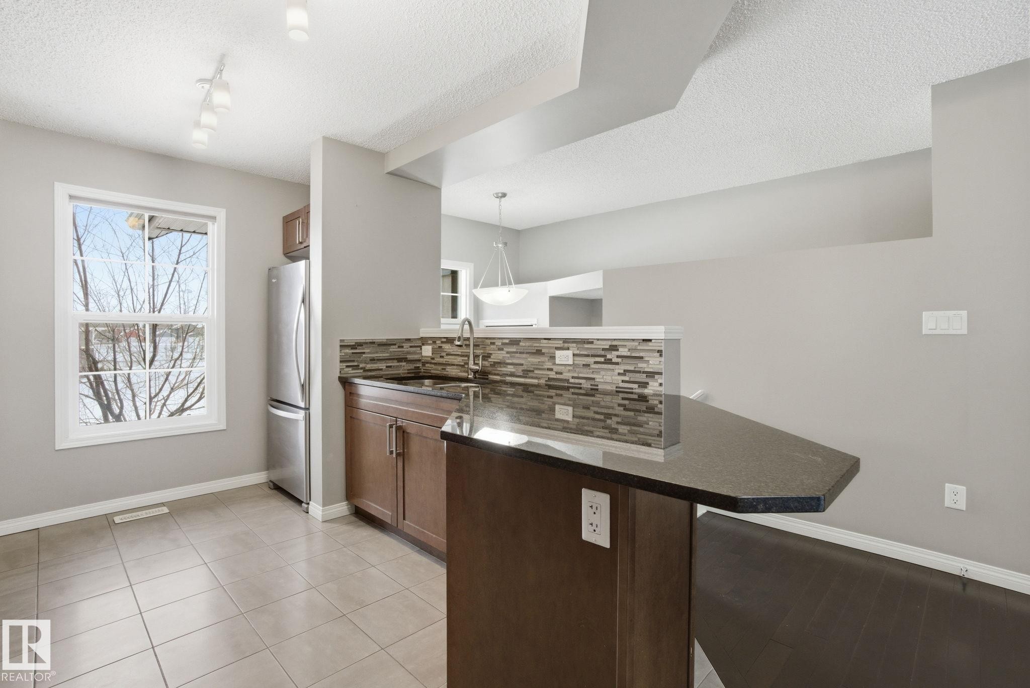 Kitchen featuring a peninsula, backsplash, freestanding refrigerator, dark stone counters, and a textured ceiling - 71 4029 Orchards Drive, Edmonton, AB - Indoor Photo Showing Kitchen