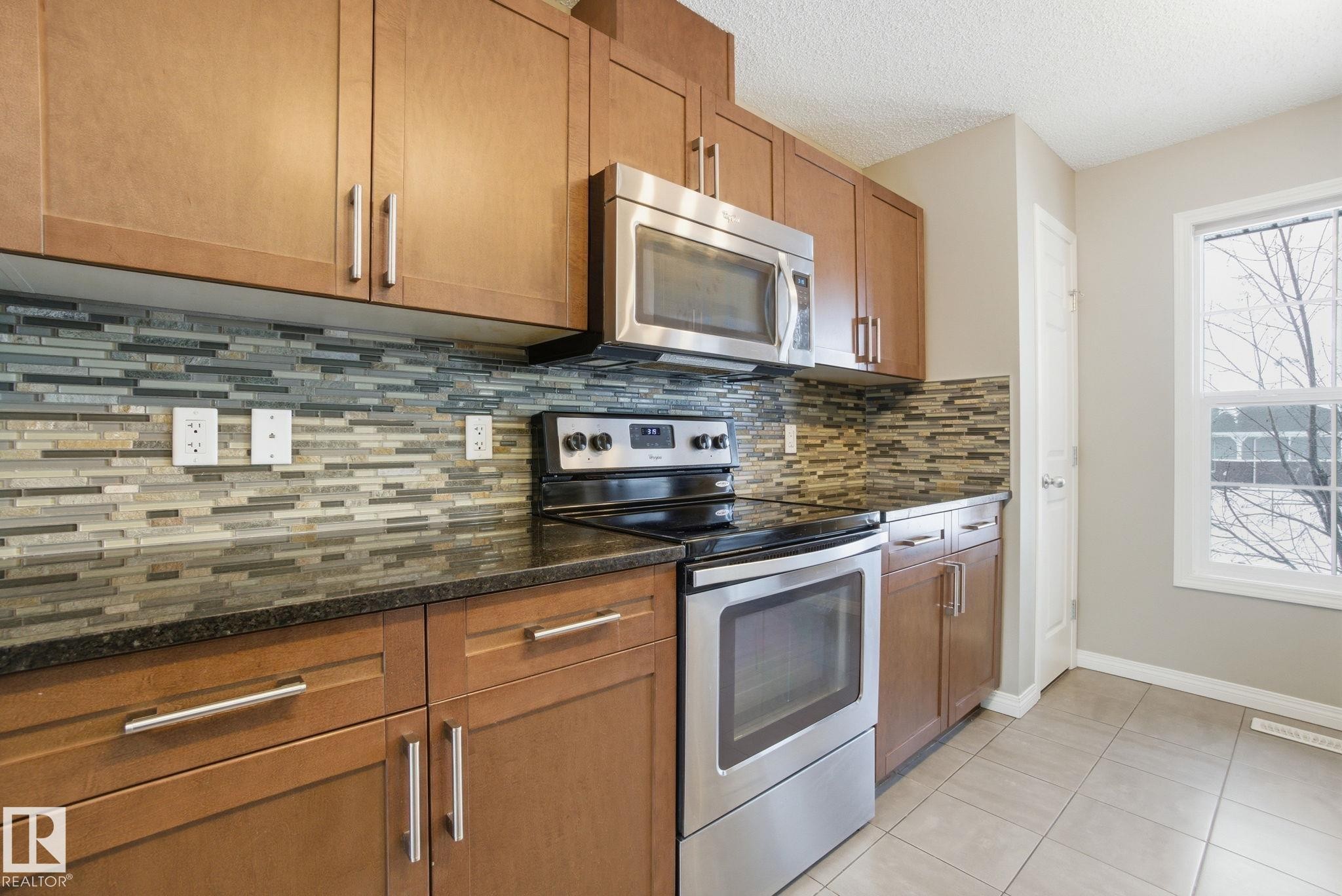 Kitchen featuring stainless steel appliances, wood finish cabinets, light tile patterned floors, dark stone counters, and a textured ceiling - 71 4029 Orchards Drive, Edmonton, AB - Indoor Photo Showing Kitchen With Upgraded Kitchen