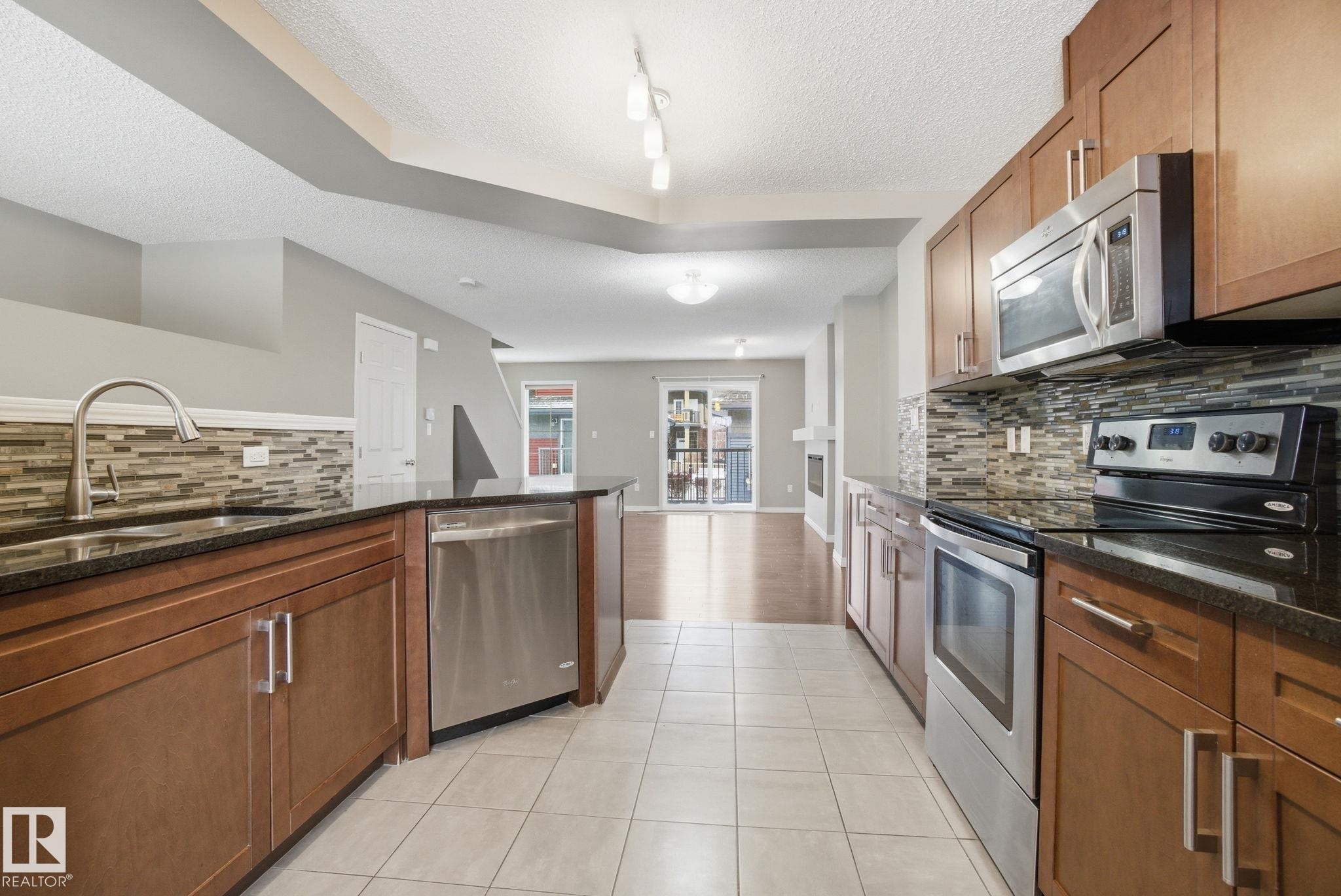 Kitchen with stainless steel appliances, dark stone countertops, a textured ceiling, wood finish cabinets, and decorative backsplash - 71 4029 Orchards Drive, Edmonton, AB - Indoor Photo Showing Kitchen With Stainless Steel Kitchen With Upgraded Kitchen