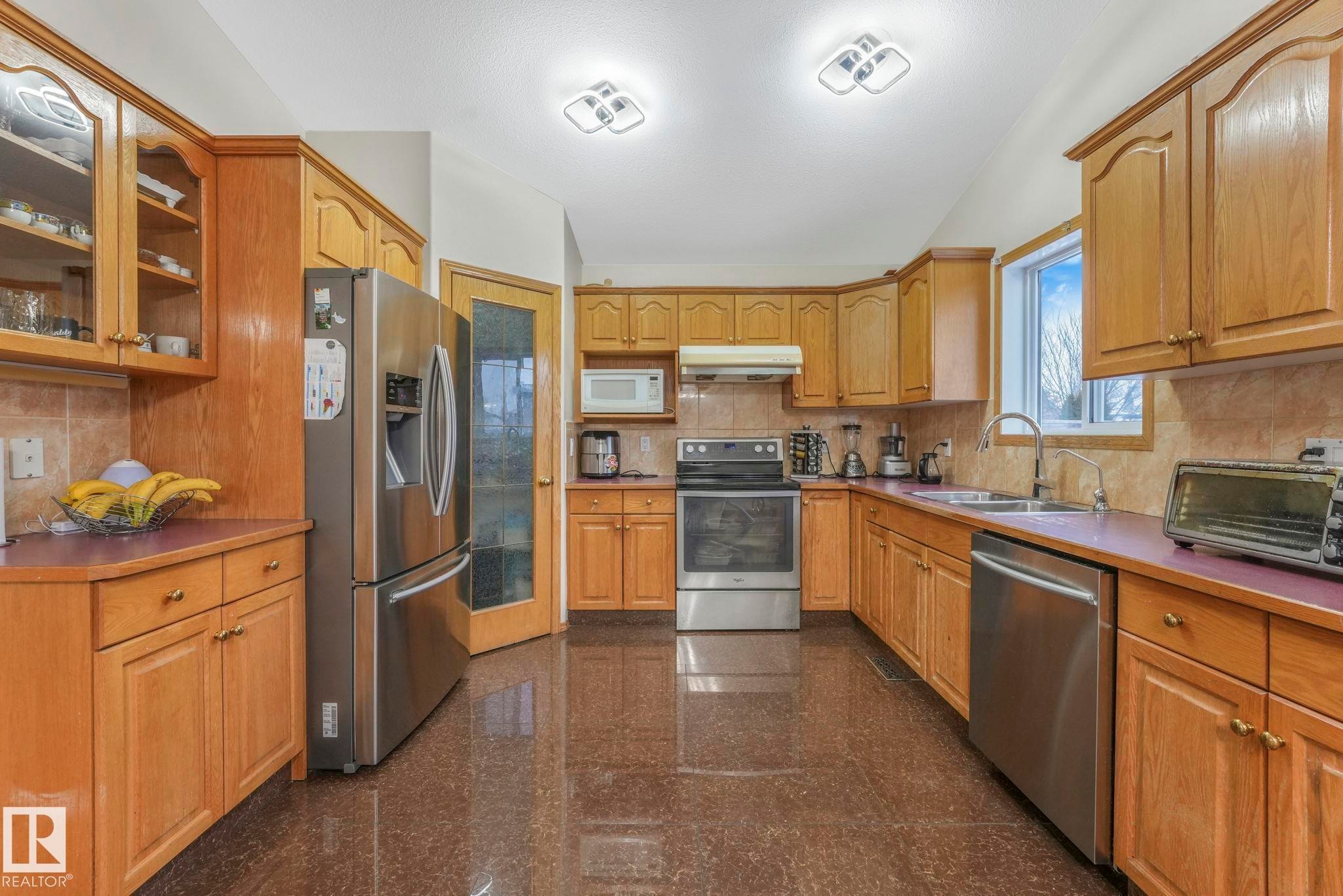 Kitchen with granite tiled floors, appliances with stainless steel finishes, glass insert cabinets, tasteful backsplash, and under cabinet range hood - 4720 150 Avenue, Edmonton, AB - Indoor Photo Showing Kitchen With Double Sink