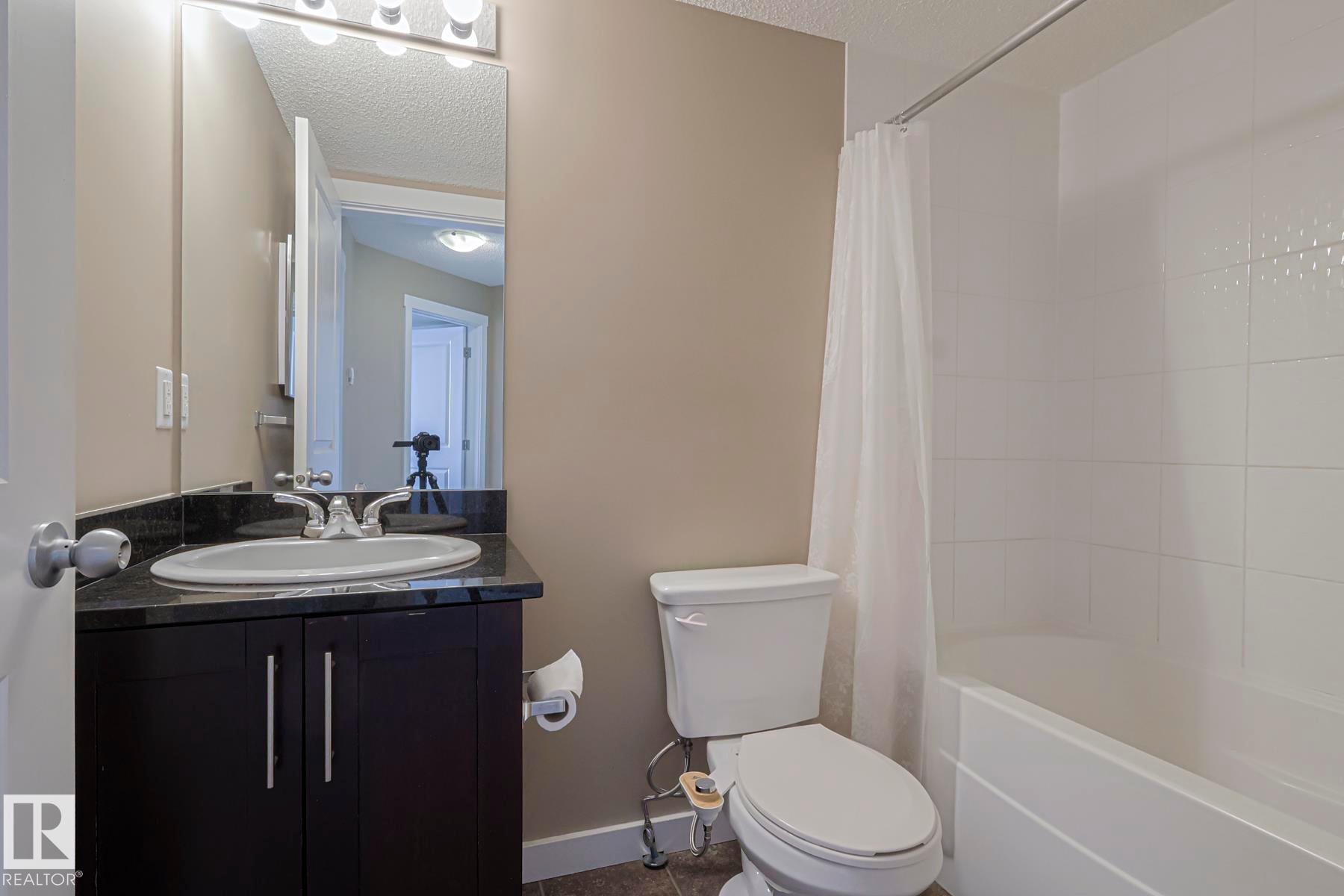 Bathroom featuring a dark wood vanity with a stone countertop and an integrated sink, a mirror with overhead lighting, and a white bathtub with a shower curtain - 313 1510 Watt Drive, Edmonton, AB - Indoor Photo Showing Bathroom
