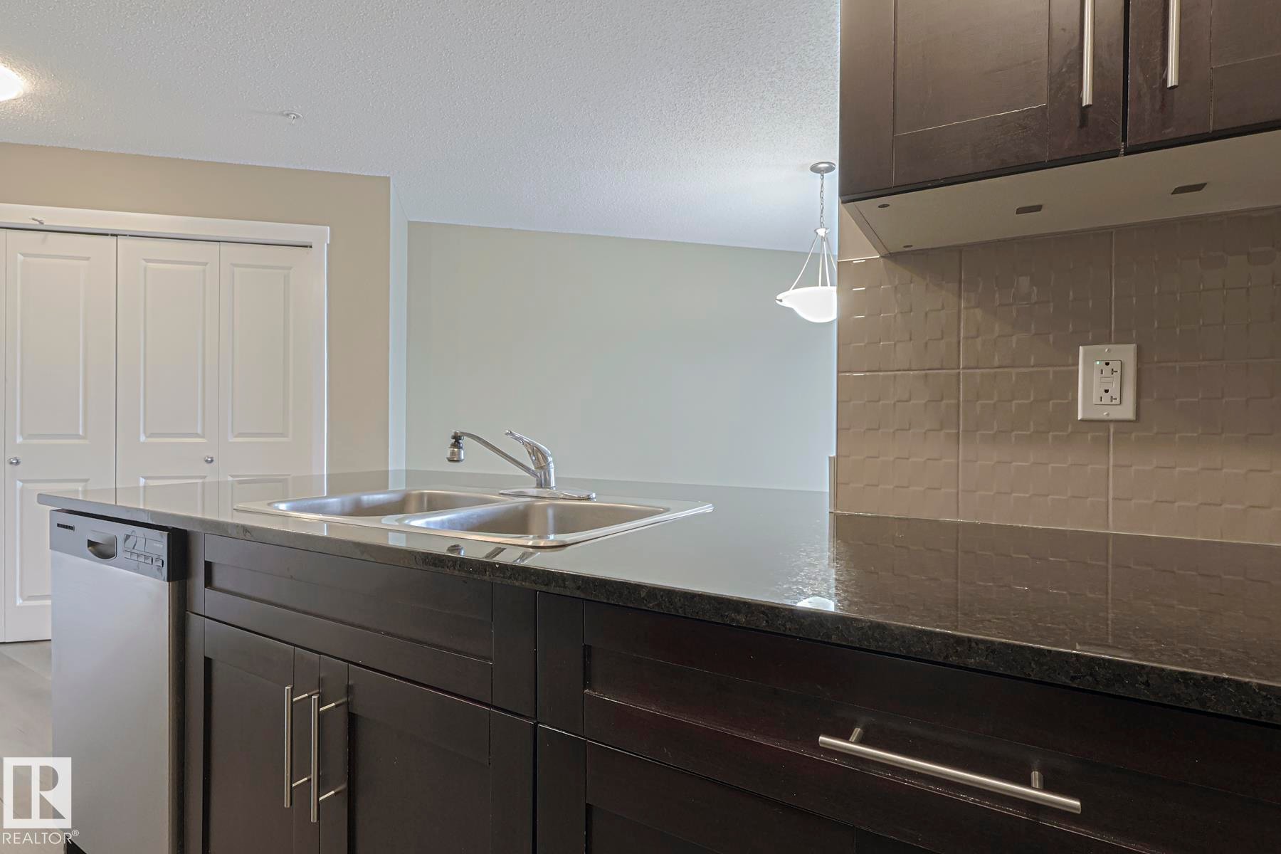 Kitchen featuring dark cabinetry, stainless steel dishwasher, double basin sink, and a tiled backsplash - 313 1510 Watt Drive, Edmonton, AB - Indoor Photo Showing Kitchen With Double Sink