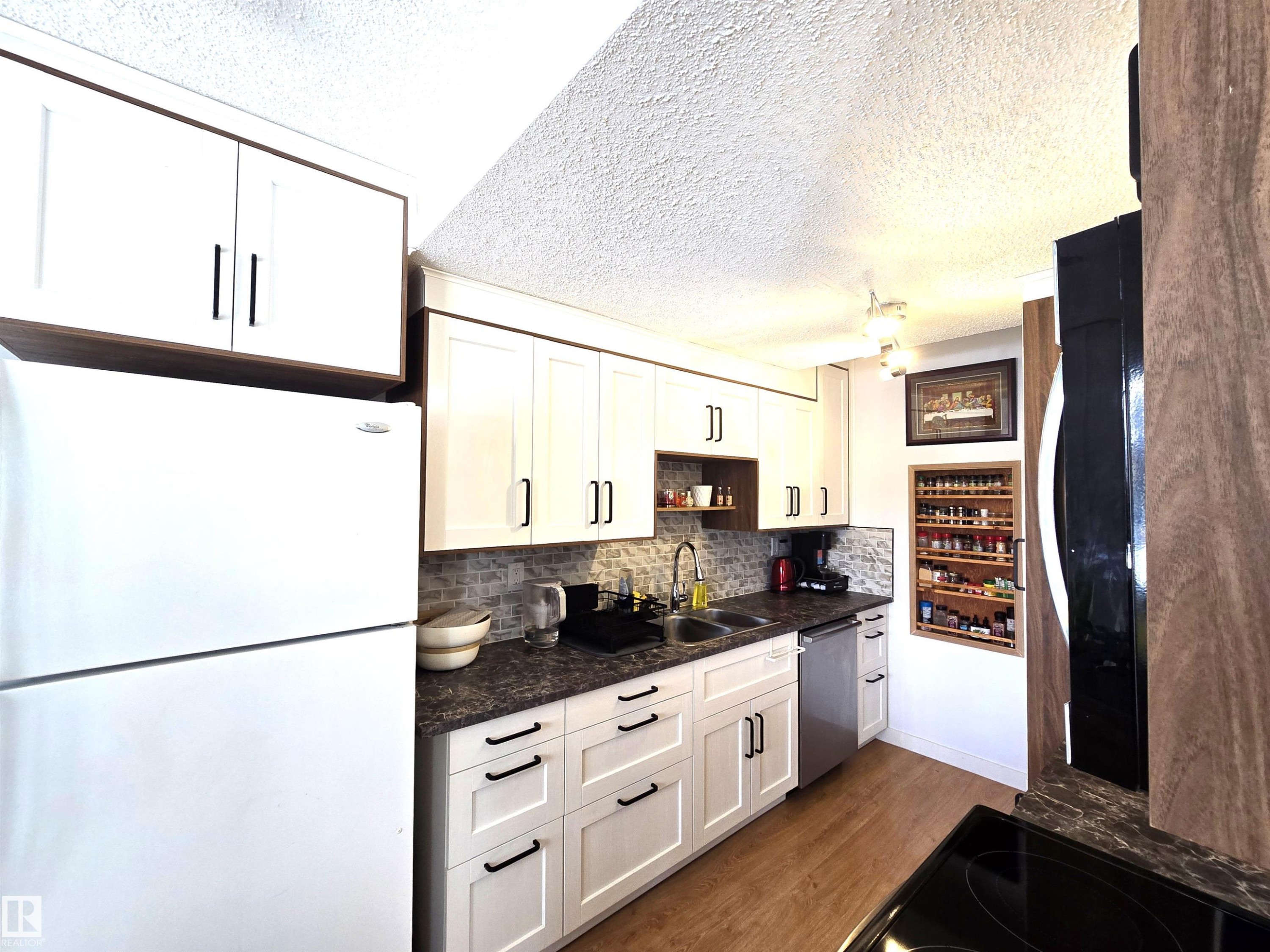 The kitchen features white cabinetry with dark hardware, dark countertops, a tile backsplash, and wood-style flooring - 80 Amberly Crest, Edmonton, AB - Indoor Photo Showing Kitchen With Double Sink