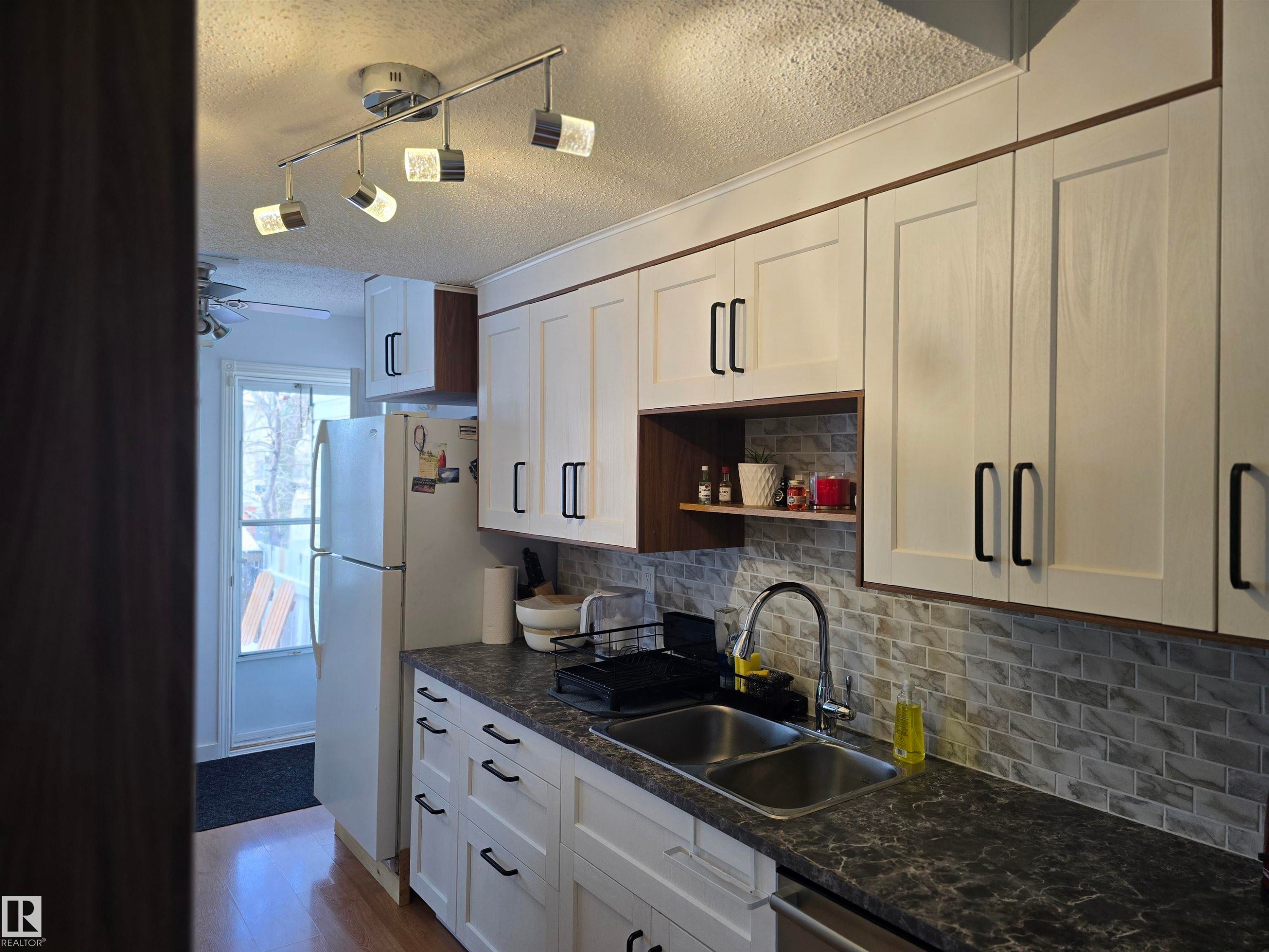 The kitchen features white cabinetry with black hardware, a dual basin stainless steel sink, a tiled backsplash, and track lighting - 80 Amberly Crest, Edmonton, AB - Indoor Photo Showing Kitchen With Double Sink