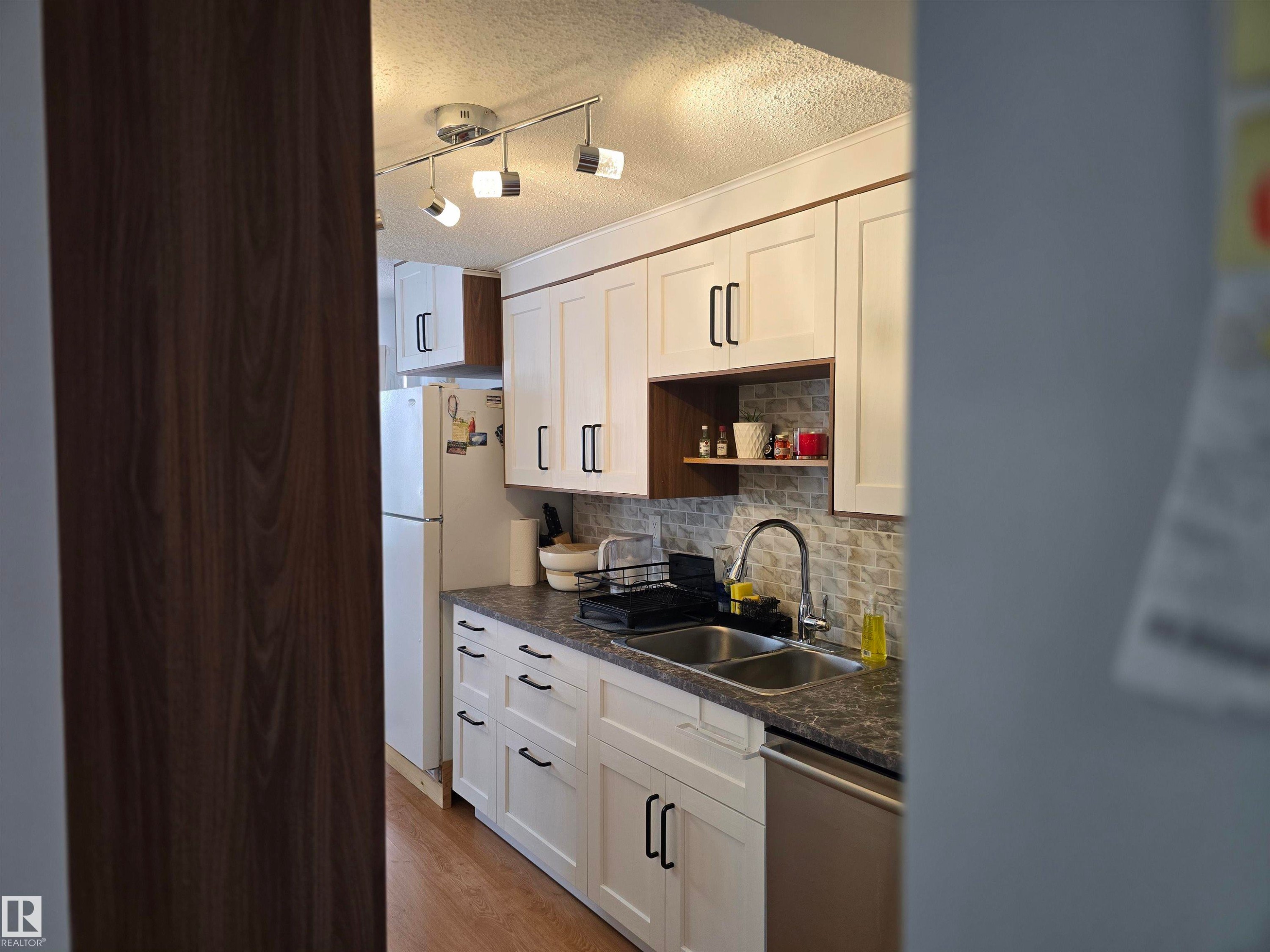 The kitchen features white cabinetry with dark hardware, a double basin sink with a gooseneck faucet, and a light-colored tile backsplash - 80 Amberly Crest, Edmonton, AB - Indoor Photo Showing Kitchen With Double Sink