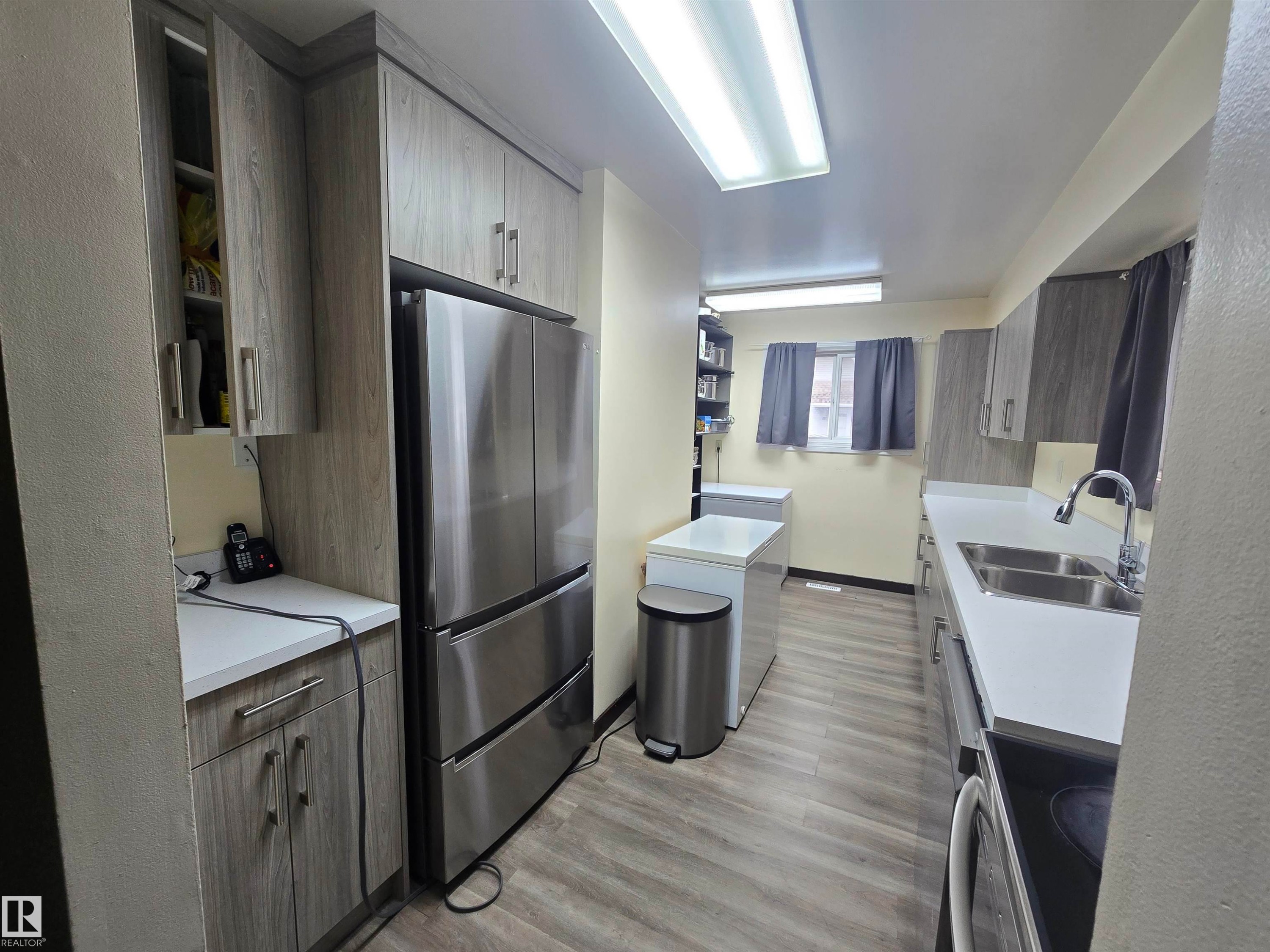 Galley kitchen featuring wood-finish cabinetry, stainless steel refrigerator, white countertops, double basin sink, and wood-finish flooring - 2 4403 Riverbend Road, Edmonton, AB