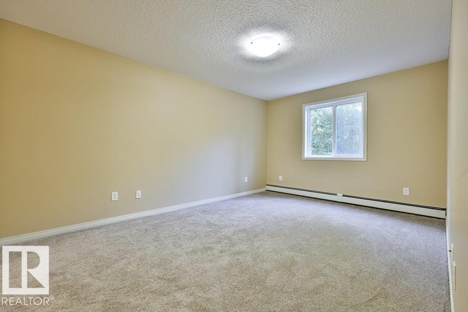 Spacious room featuring neutral carpeting, light beige walls, a white window frame, and baseboard heating - #107 4903 47 Av, Stony Plain, AB - Indoor Photo Showing Other Room