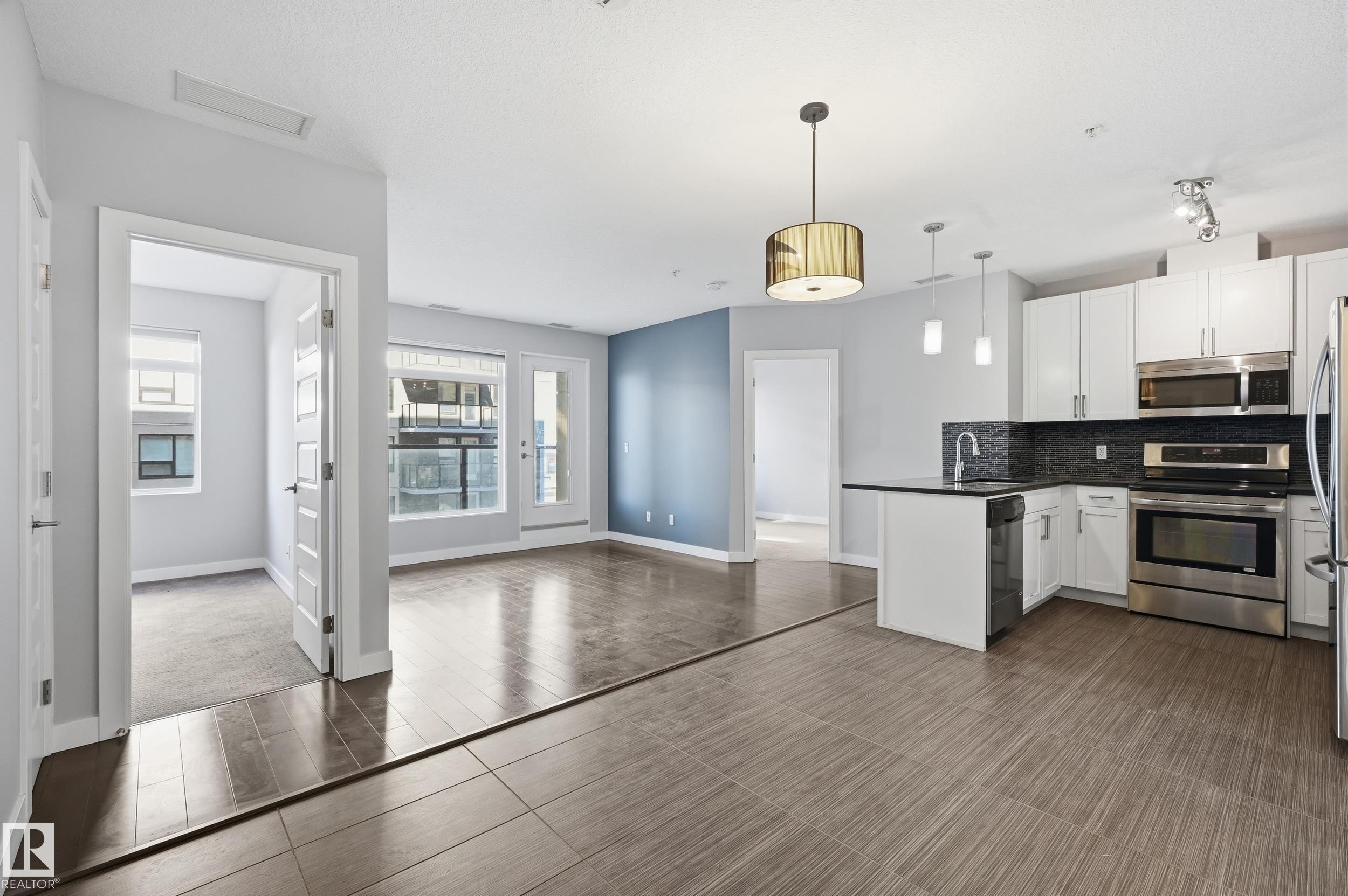 Kitchen with stainless steel appliances, backsplash, white cabinetry, decorative light fixtures, and open floor plan - 511 5151 Windermere Boulevard, Edmonton, AB - Indoor Photo Showing Kitchen