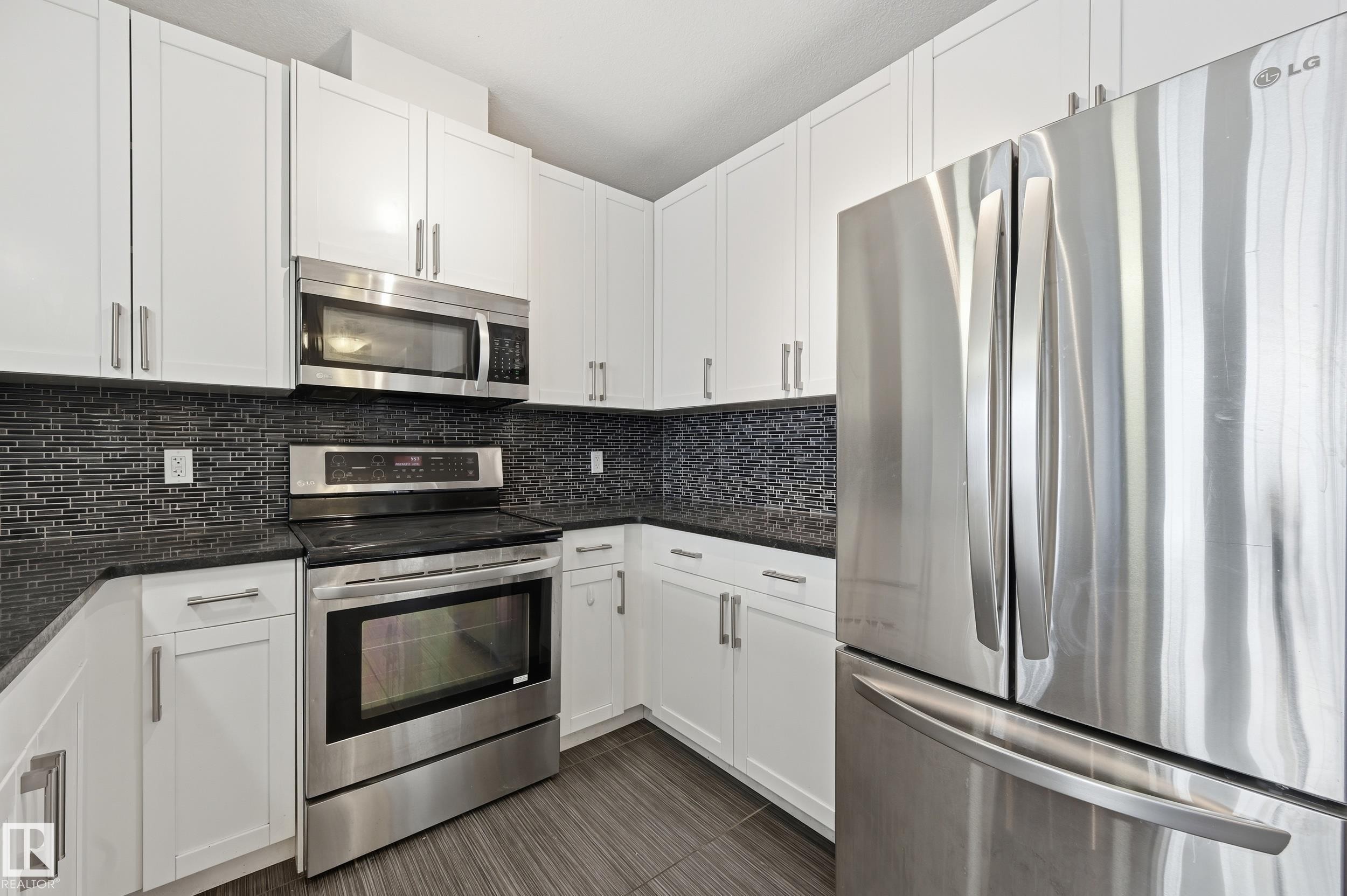 Kitchen featuring stainless steel appliances, white cabinetry, decorative backsplash, and dark stone counters - 511 5151 Windermere Boulevard, Edmonton, AB - Indoor Photo Showing Kitchen With Upgraded Kitchen