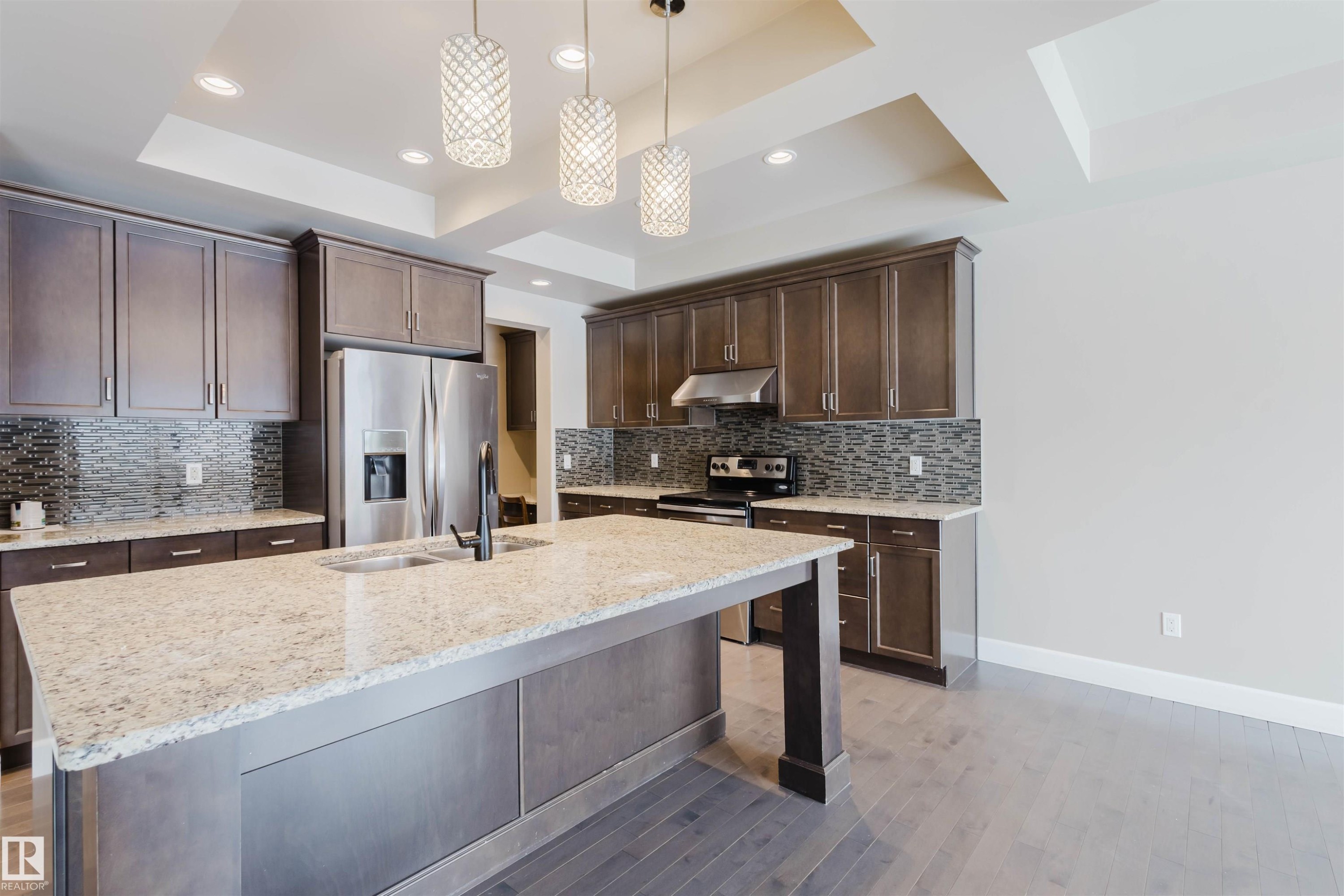 Kitchen with dark wood finish cabinets, light stone counters, stainless steel appliances, a tray ceiling, and an island with sink - 1957 Ainslie Link, Edmonton, AB - Indoor Photo Showing Kitchen With Stainless Steel Kitchen With Double Sink With Upgraded Kitchen