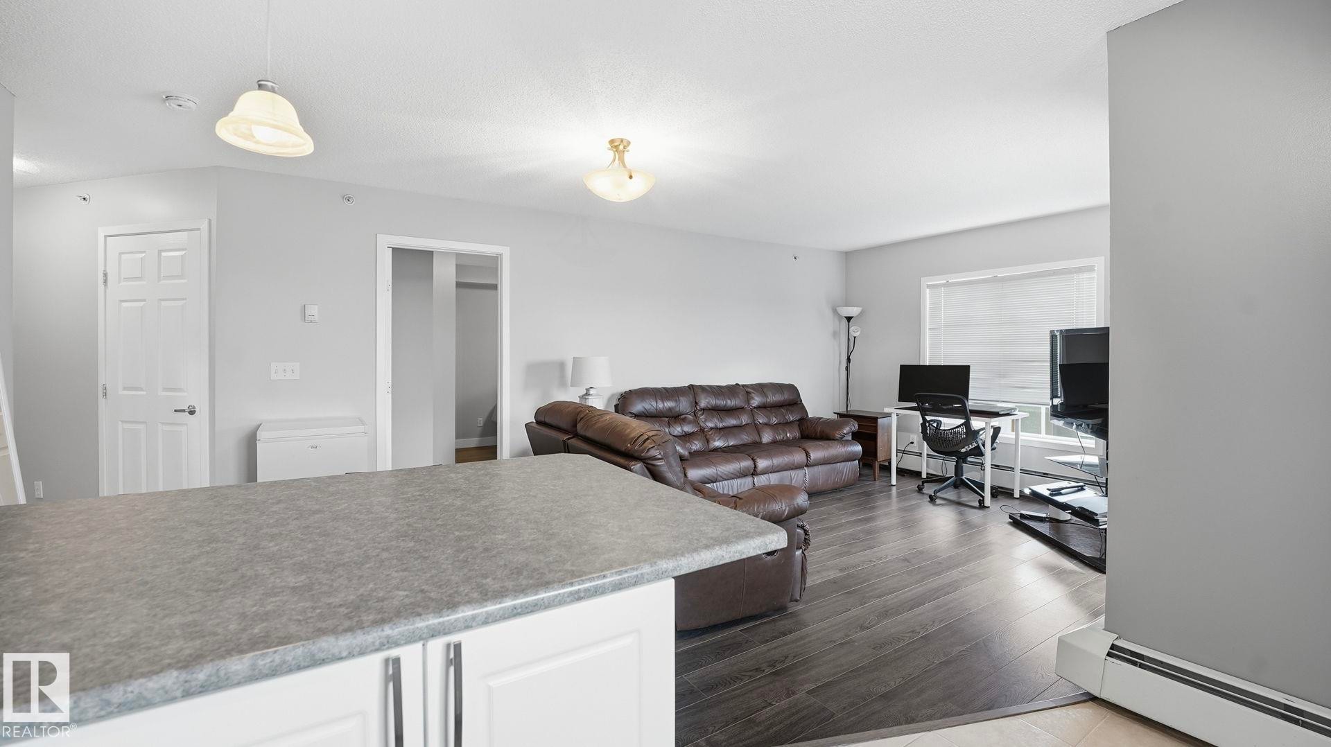 Living room with a baseboard radiator, an office area, and dark wood-style flooring - 401 636 Mcallister Loop, Edmonton, AB - Indoor