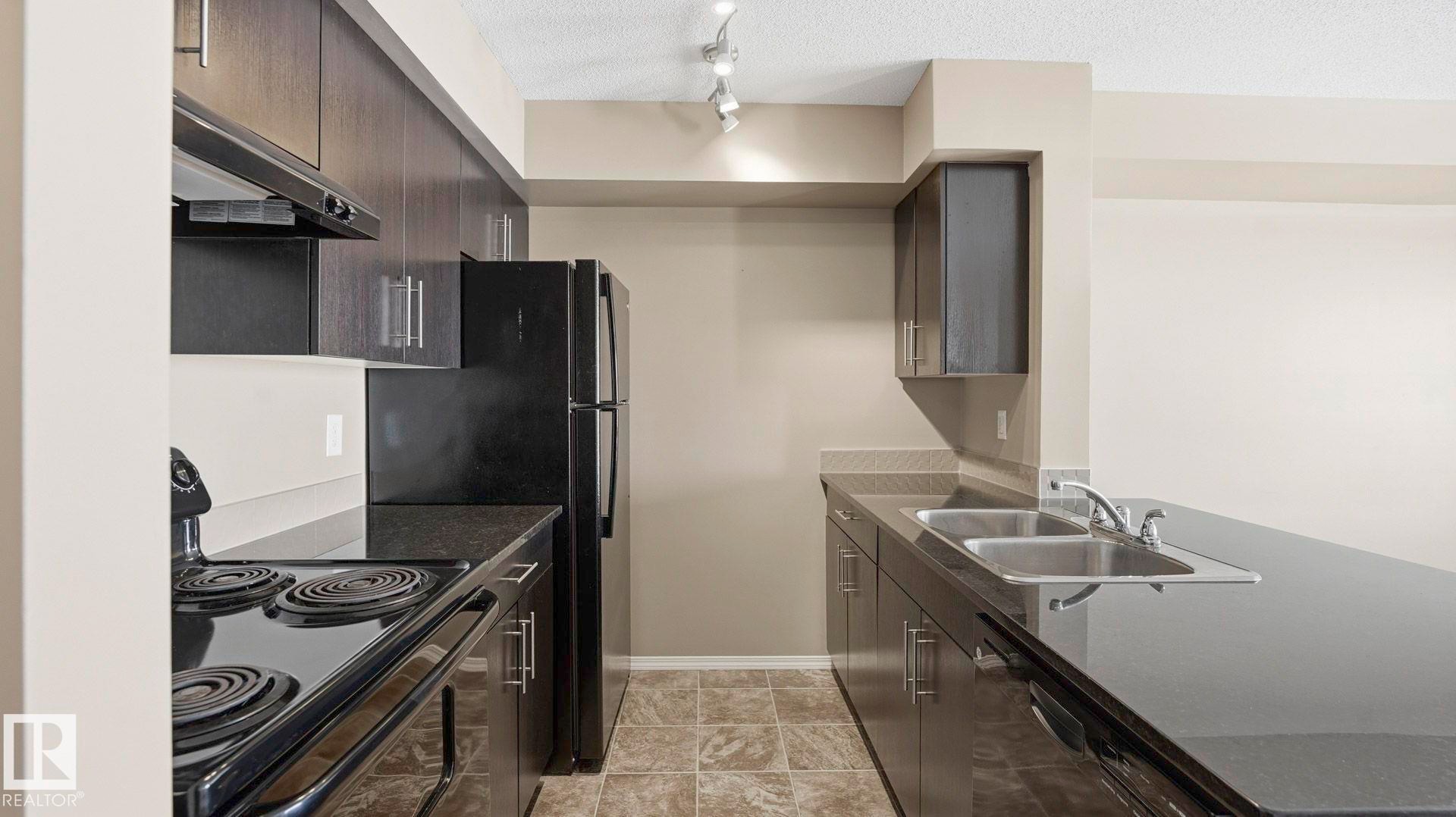 Kitchen with black appliances, dark wood finish cabinetry, a textured ceiling, dark stone countertops, and a peninsula - 319 274 Mcconachie Drive, Edmonton, AB - Indoor Photo Showing Kitchen With Double Sink