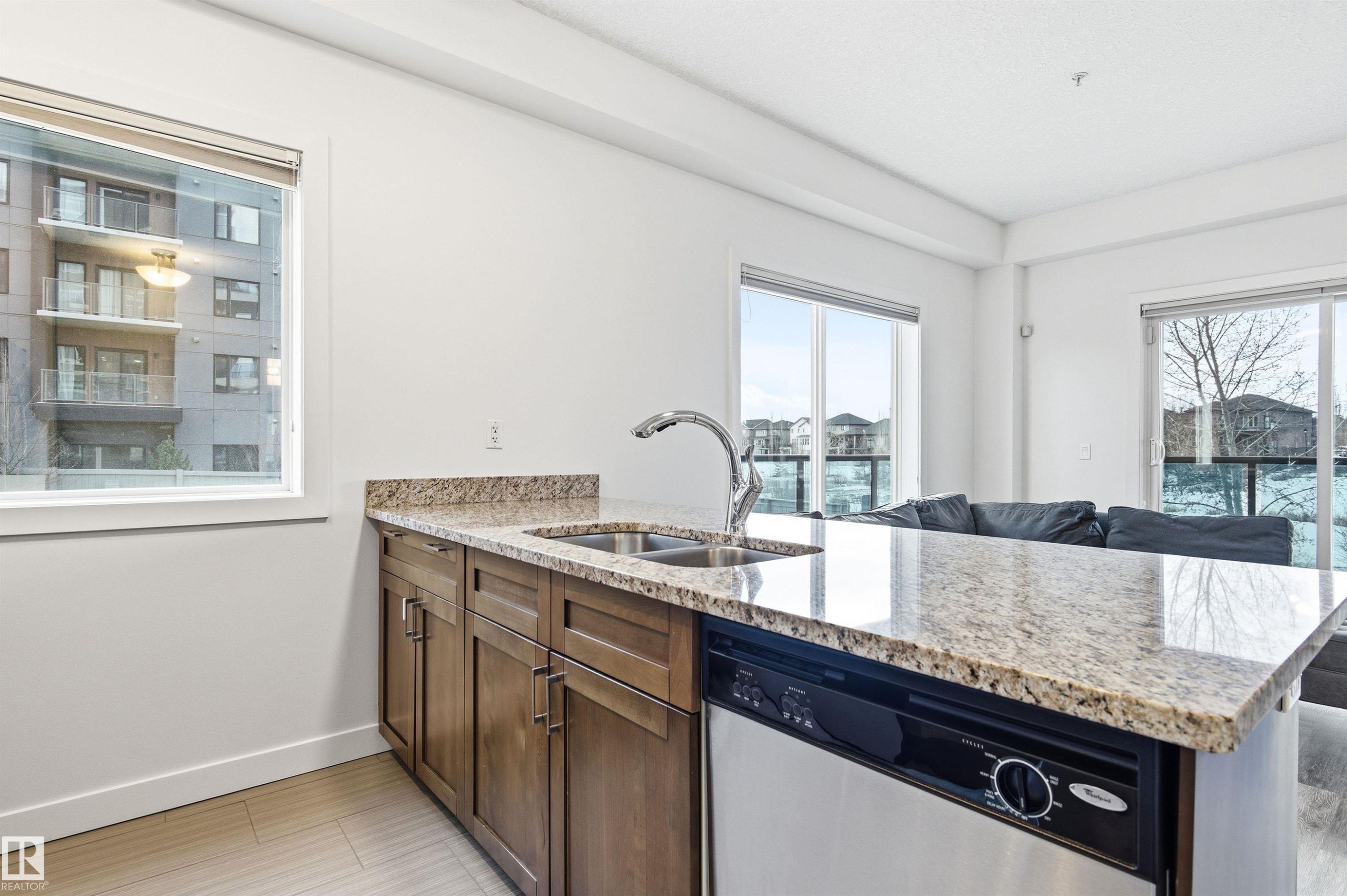 Kitchen with a peninsula, light stone counters, stainless steel dishwasher, open floor plan, and wood finish cabinets - 106 1238 Windermere Way, Edmonton, AB - Indoor Photo Showing Kitchen With Double Sink