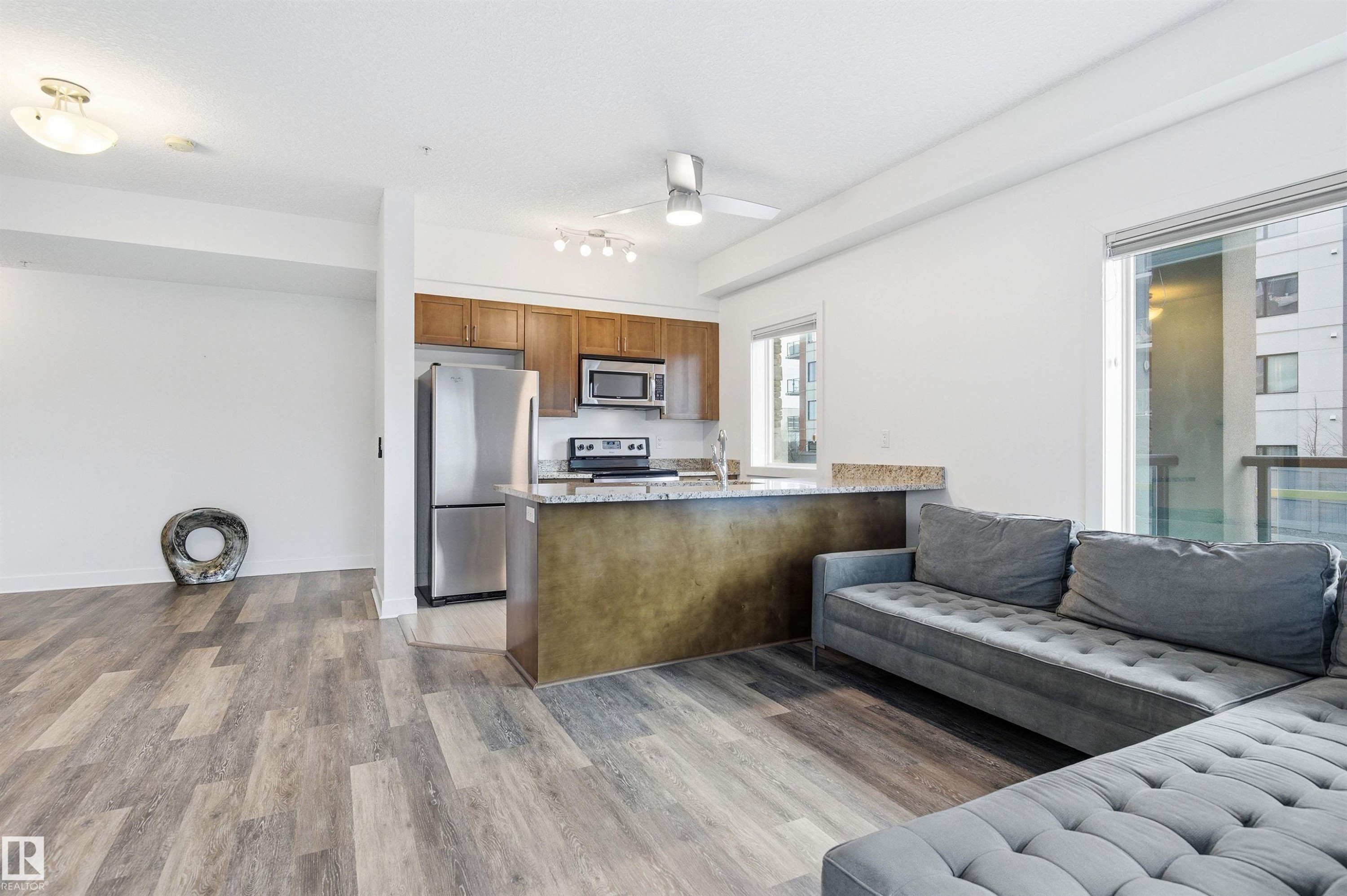 Kitchen with open floor plan, a peninsula, stainless steel appliances, light stone counters, and light wood-type flooring - 106 1238 Windermere Way, Edmonton, AB - Indoor