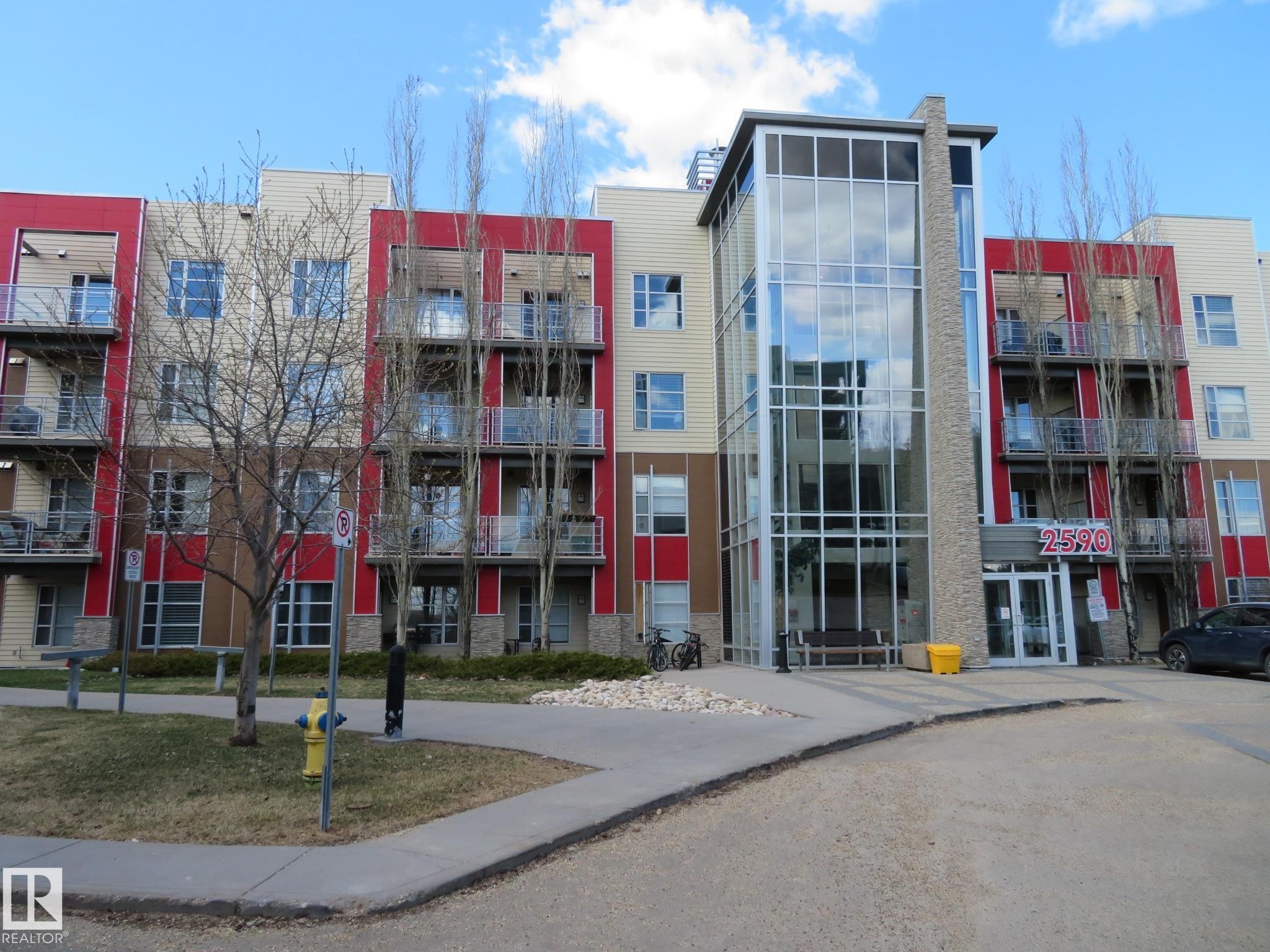 Contemporary building exterior featuring a multi-story glass entryway, red and tan facade panels, multiple private balconies with glass railings, and stone-finish detailing - 415 2590 Anderson Way, Edmonton, AB - Outdoor With Facade