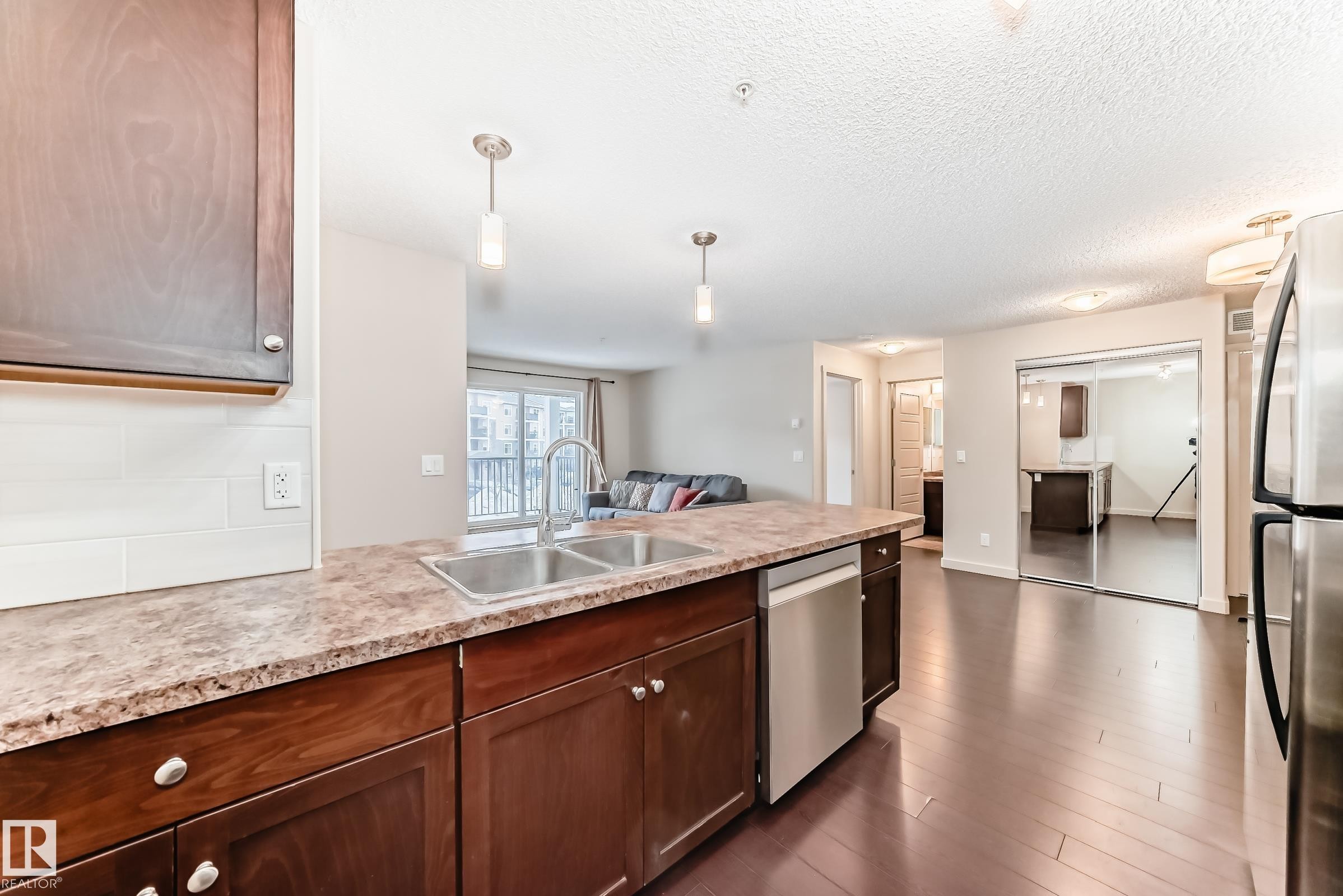 Kitchen featuring stainless steel appliances, decorative light fixtures, a peninsula, open floor plan, and dark wood-type flooring - 229 6076 Schonsee Way, Edmonton, AB - Indoor Photo Showing Kitchen With Stainless Steel Kitchen With Double Sink