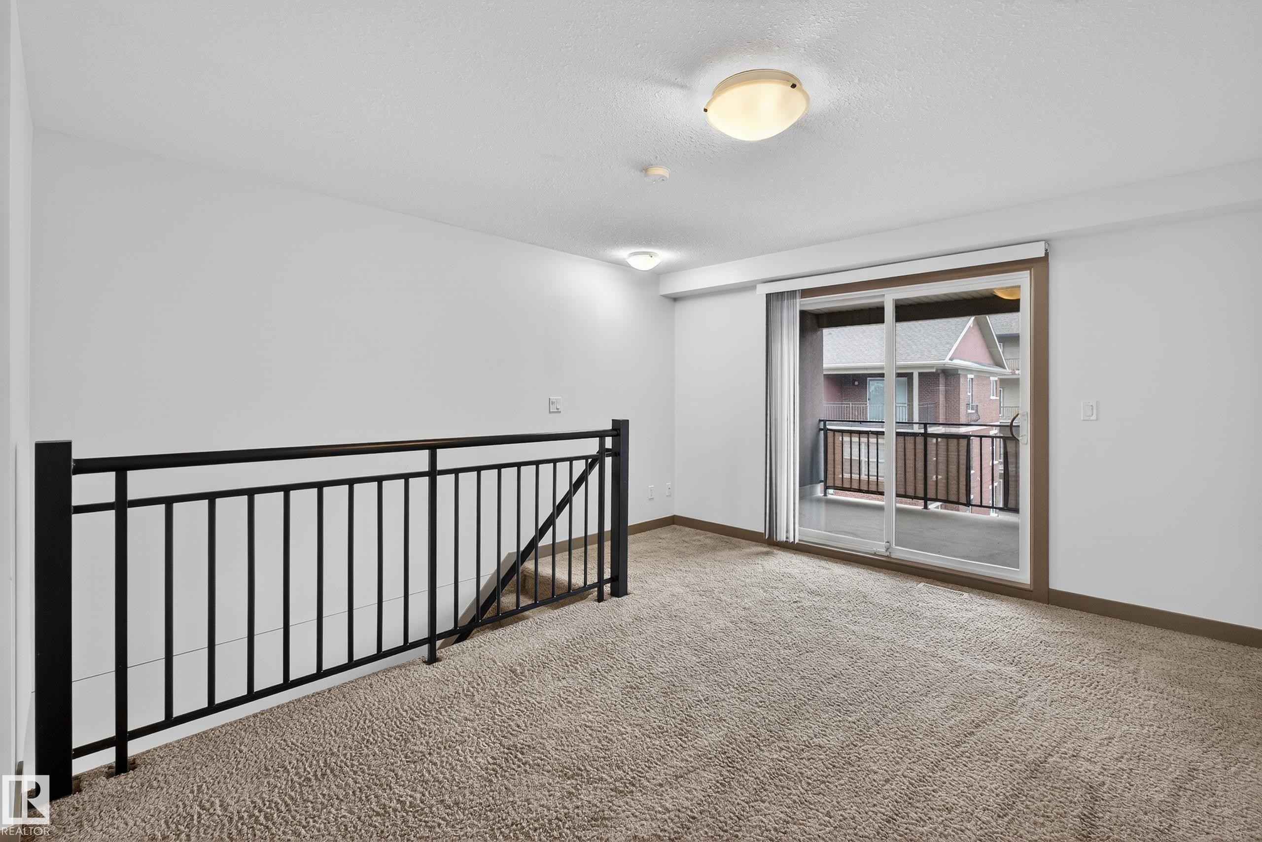 This inviting space features light-colored carpeting, a black metal railing, and a sliding glass door providing access to a balcony - 1649 Cunningham Way, Edmonton, AB - Indoor Photo Showing Other Room