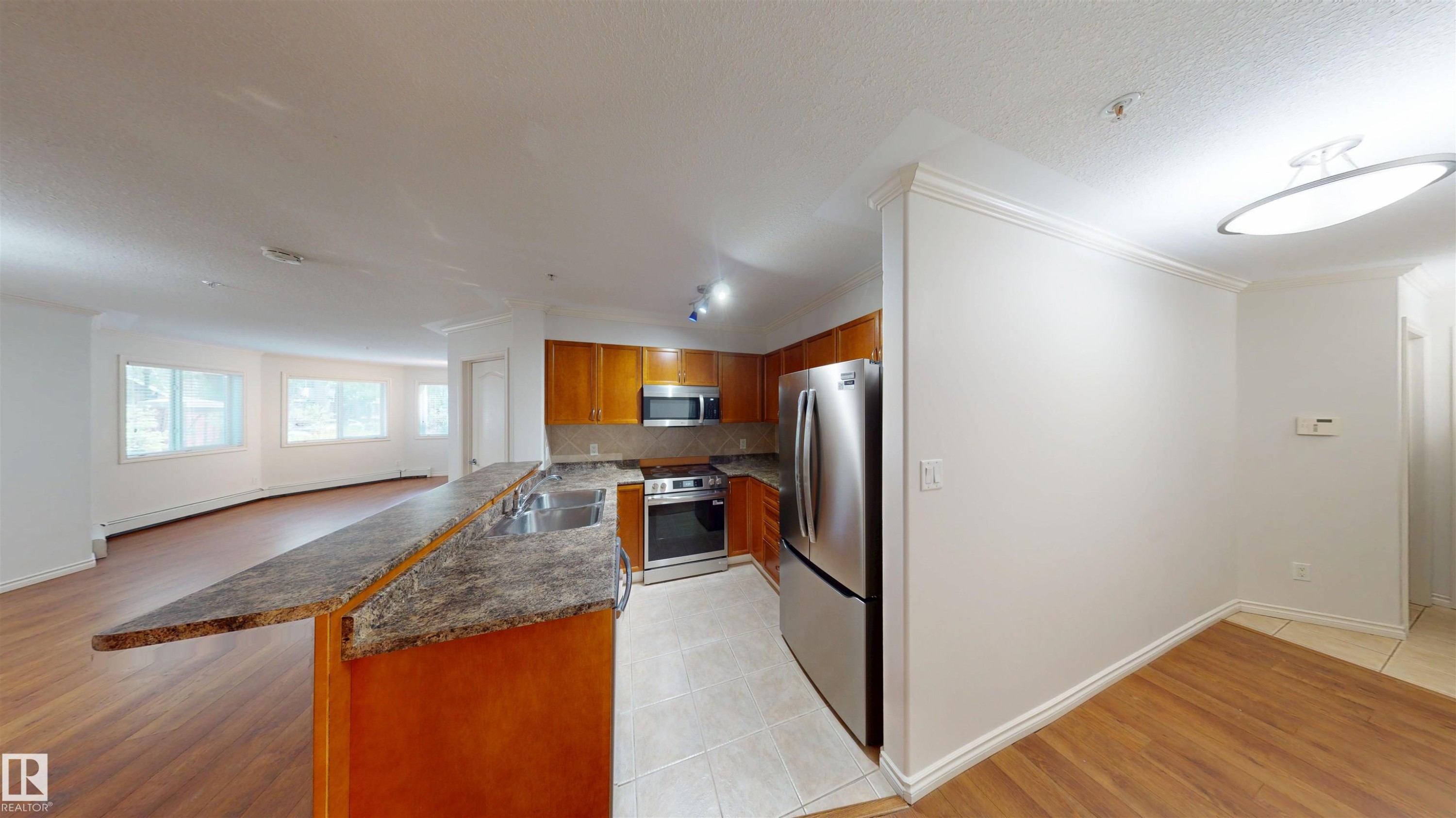 Kitchen featuring wood finish cabinets, stainless steel appliances, a peninsula, a baseboard heating unit, and light tile patterned floors - 115 9938 104 St, Edmonton, AB - Indoor Photo Showing Kitchen With Double Sink