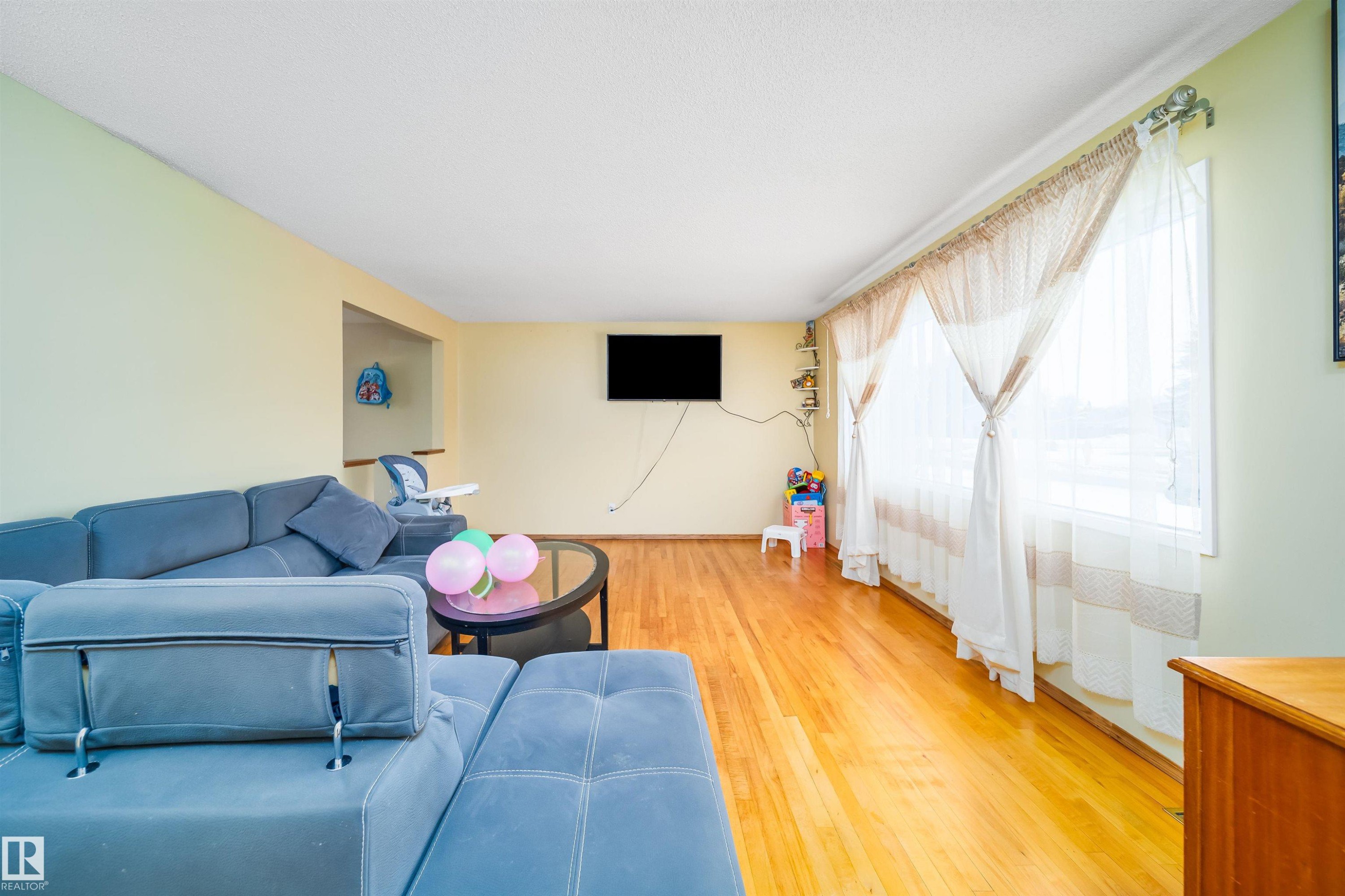 Living area featuring hardwood flooring, light-colored walls, and a large window with sheer curtains - 7911 145 Avenue Nw, Edmonton, AB - Indoor Photo Showing Living Room