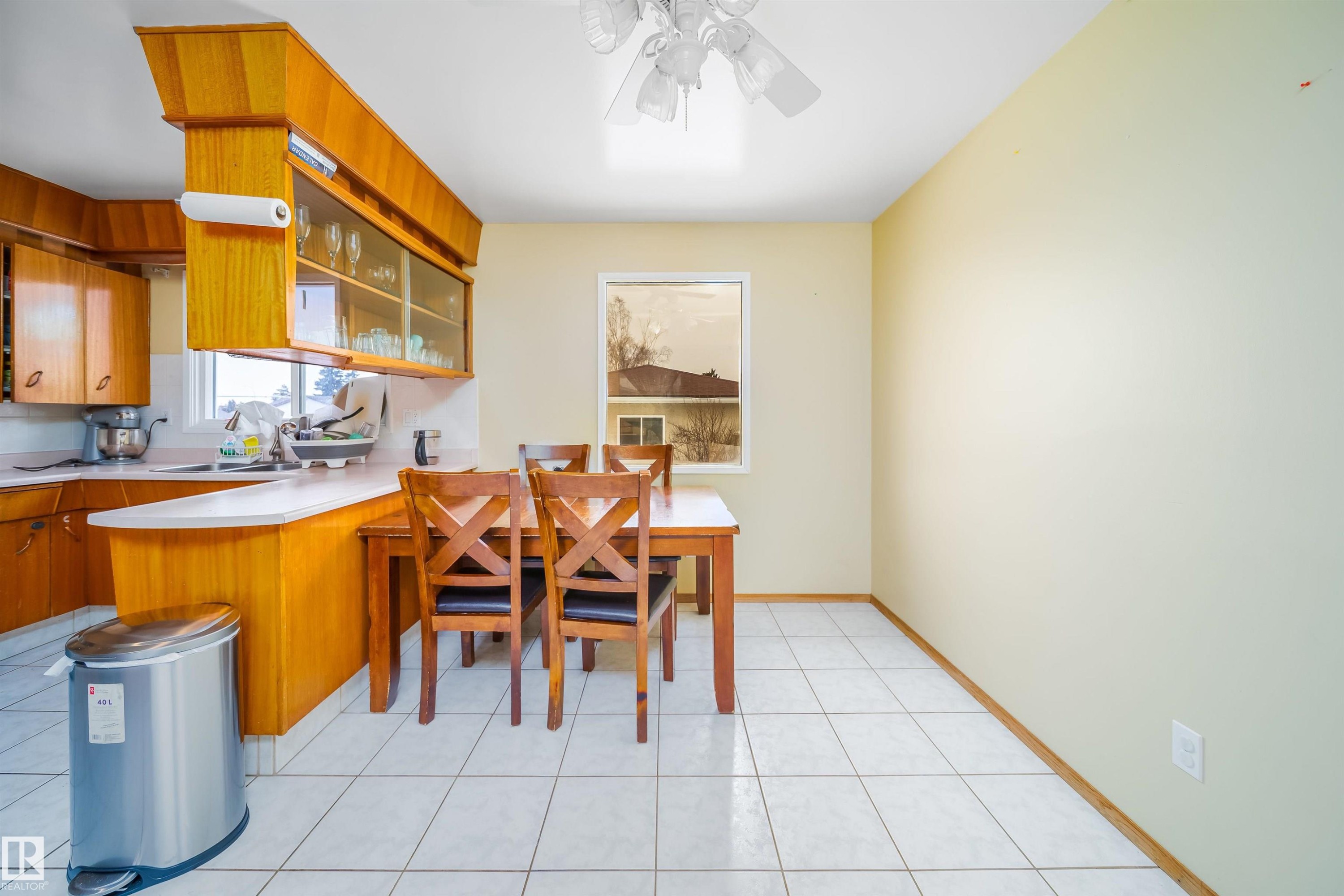 Kitchen and dining area featuring light-colored tile flooring, a window, and wooden cabinetry with glass-fronted upper cabinets - 7911 145 Avenue Nw, Edmonton, AB - Indoor