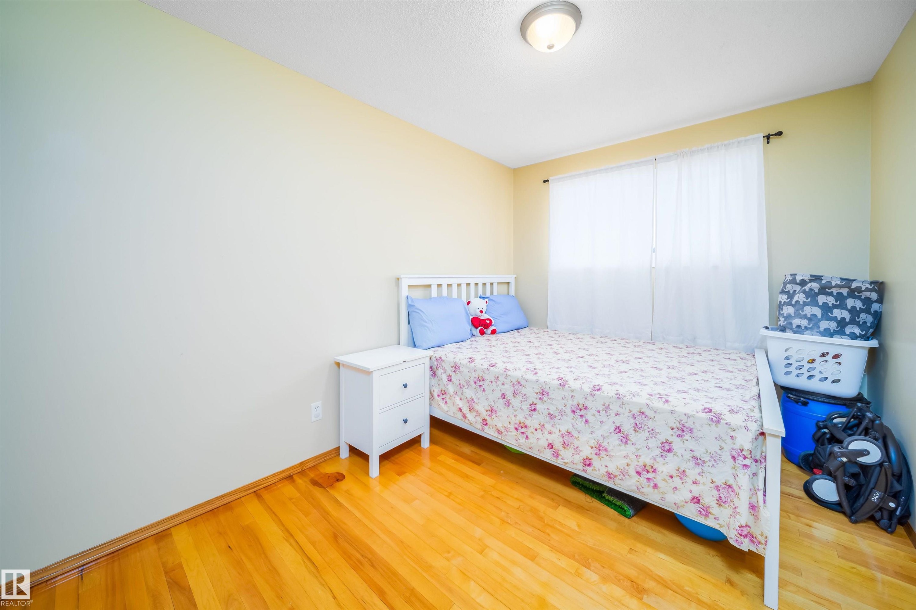 This room features hardwood flooring, a white bed frame, and a window with white curtains - 7911 145 Avenue Nw, Edmonton, AB - Indoor Photo Showing Bedroom